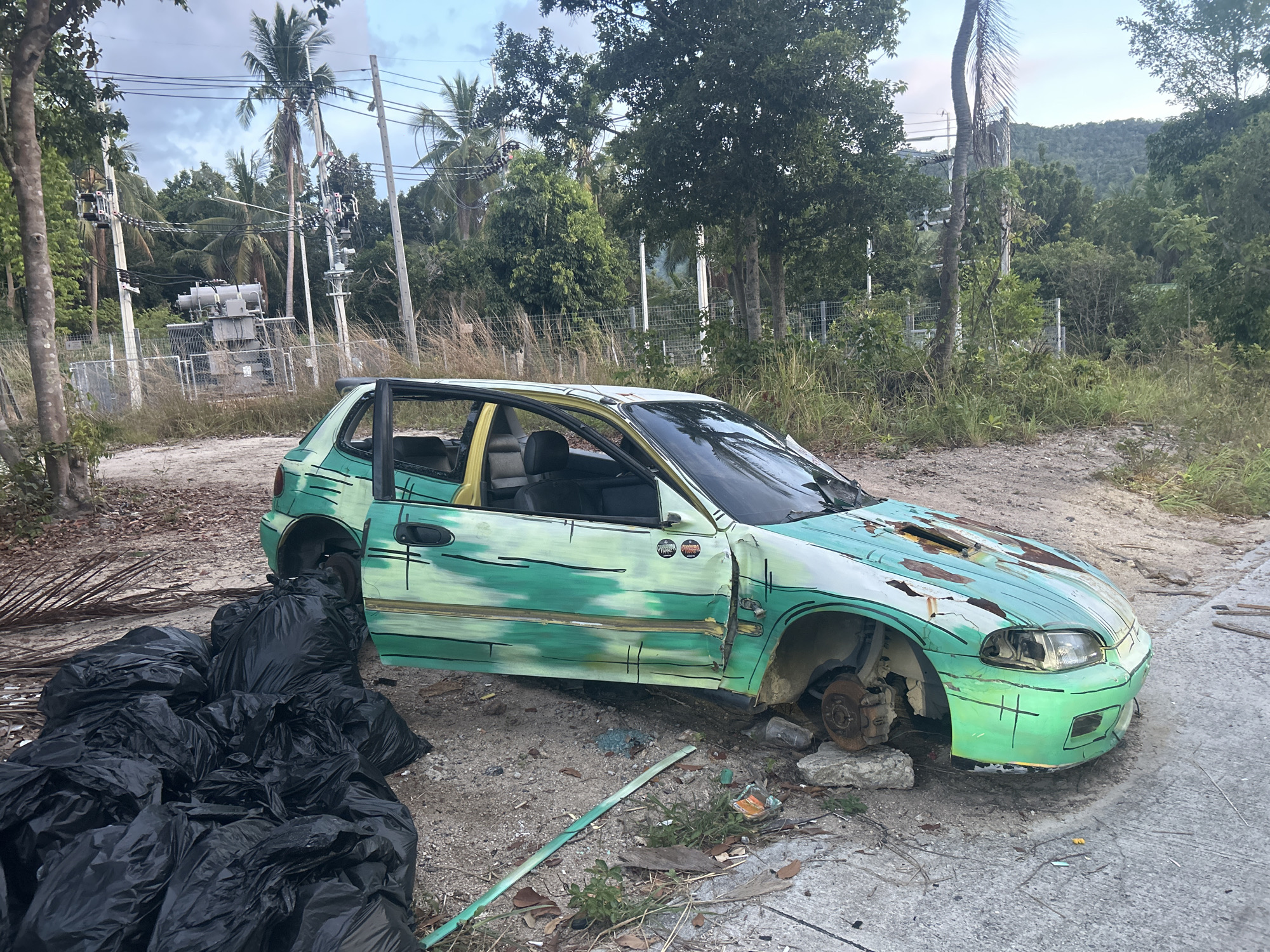 An abandoned, heavily damaged, green and white Honda Civic is on the side of a road in Baan Wang Ta Kien, Thailand. The car's paint is peeling, and rust is visible on the hood. The front passenger-side wheel is missing, exposing the brake assembly. The driver-side door is open. There are several large, black trash bags piled near the car. The background features lush tropical vegetation, including palm trees and power lines. A fenced-in electrical substation is visible in the distance. The sky is overcast.