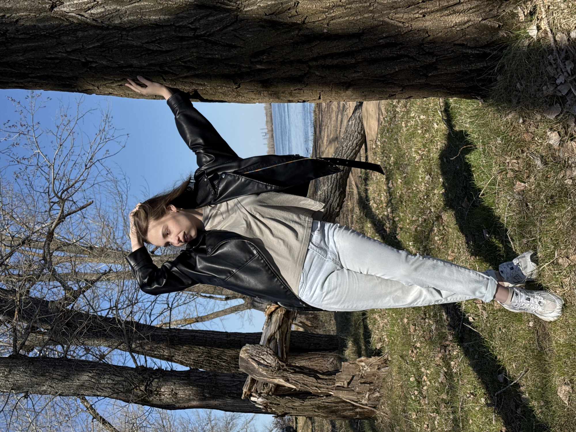 A young woman poses next to a fallen tree on a sunny day. She wears a black leather jacket, a beige t-shirt, light-wash jeans, and white sneakers. She has one hand on the tree and another on her head. In the background is green grass and leafless trees. A glimpse of a body of water can be seen in the distance. The photo was taken in Noviy, Russia.