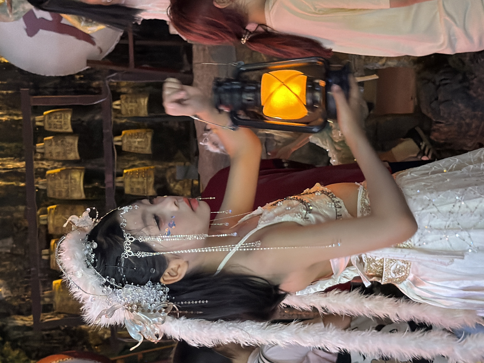 The image shows a young woman in traditional Chinese attire, possibly for a performance or cultural event, set against a dimly lit backdrop. She is wearing an ornate white dress adorned with pearls and intricate gold embroidery. A feathered headdress with crystals and dangling silver chains accents her dark hair. She holds a lit oil lantern in her raised right hand, its warm glow illuminating her face and the surrounding area. Her eyes are closed, and her lips are slightly parted, suggesting a moment of serene contemplation or dramatic expression.
The background reveals a rustic setting with rows of what appear to be ancient bronze bells mounted on a wooden structure. The lighting is atmospheric, with the lantern and ambient light creating shadows and highlighting the textures of the scene. The overall mood is one of mystery and tradition, with a touch of enchantment due to the woman's elaborate costume and the glow of the lantern. The time of day appears to be evening or night, given the darkness and the reliance on artificial light. The location cues, such as the bells and the overall aesthetic, suggest a setting related to traditional Chinese culture, possibly in a historical or ceremonial space within Yuzhong District, China. There is no visible text in the image.