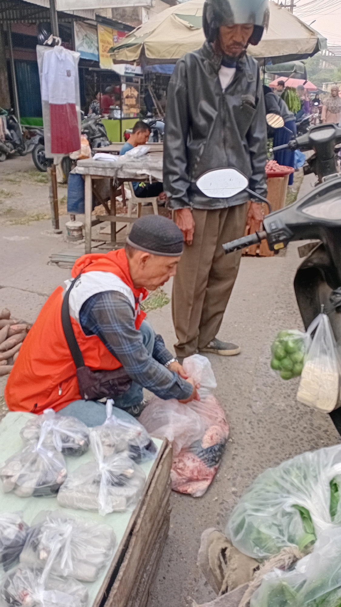 This image depicts an outdoor market scene in Jambi, Indonesia. A man wearing an orange vest over a plaid shirt and a black skullcap is kneeling, arranging items in plastic bags. Next to him, on his left, is a wooden crate filled with bagged goods. To his right, a larger plastic bag contains raw meat. In the background, another man wearing a black jacket and helmet stands, looking down. The market appears to be located on a paved road with various stalls and parked motorcycles visible. One stall displays school uniforms, suggesting proximity to educational institutions or a market catering to students. The overall atmosphere is that of a typical Indonesian street market.