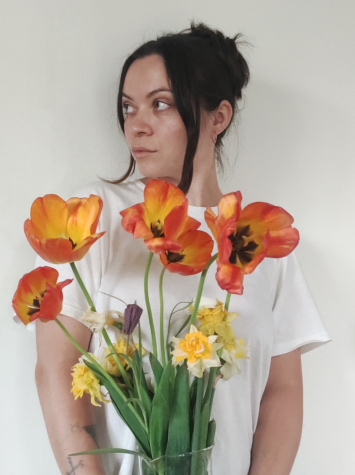 A woman with dark hair in a messy bun, wearing a plain white t-shirt, holds a bouquet of flowers. The flowers are mainly tulips in shades of orange and yellow, and some daffodils. The woman is looking to her left. Her arms are holding the bouquet. The setting seems to be indoors against a white wall. The city or specific location are not discernible.