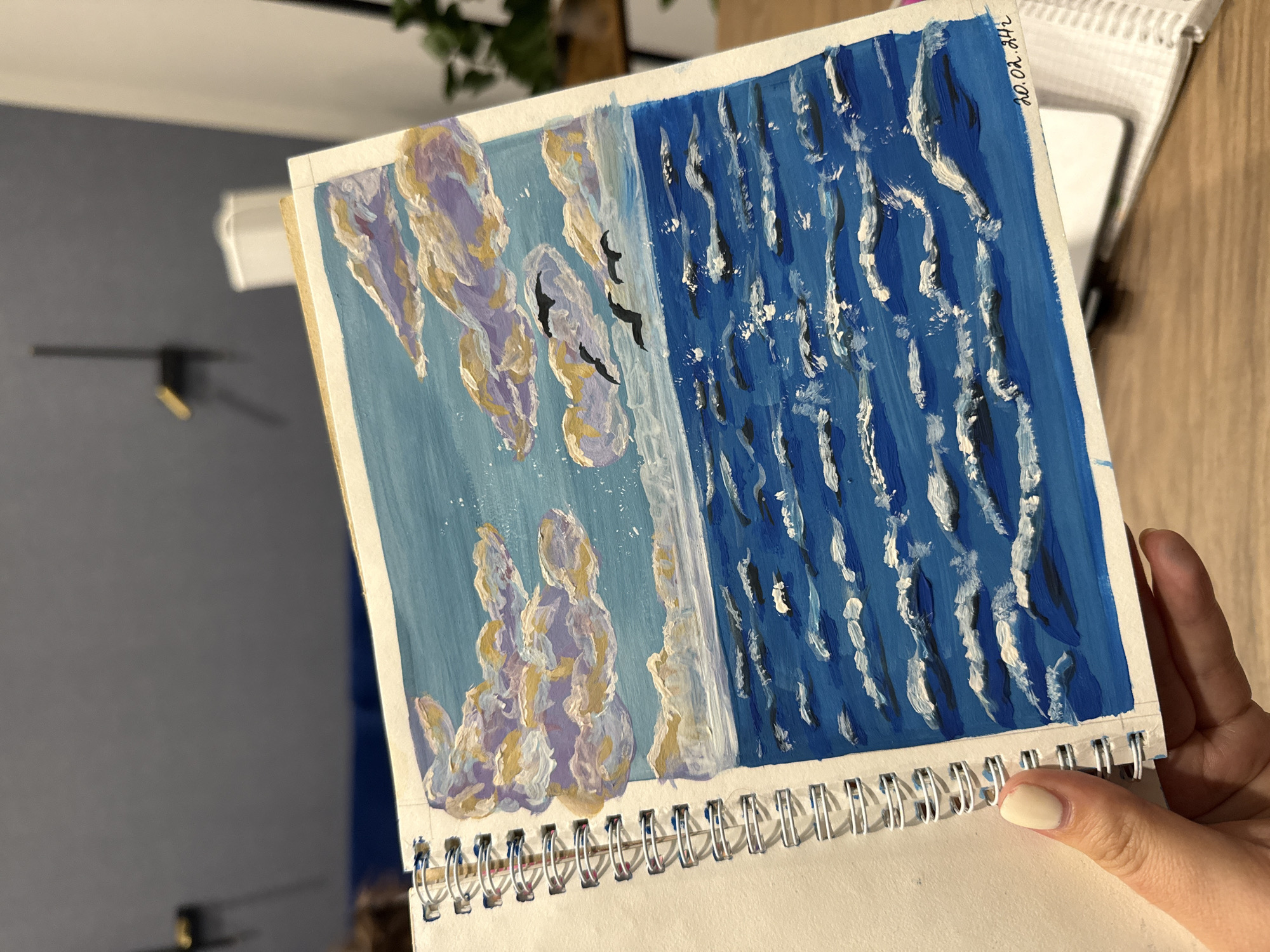 A hand is holding a spiral-bound sketchbook open to a painting of a seascape. The painting depicts a blue sky filled with fluffy clouds in shades of white, yellow, and purple. Three black birds are flying across the sky. Below the sky is a choppy blue ocean with white-capped waves. The bottom right corner of the painting has a handwritten date: "20.02.24". The background of the image shows a blurred office environment with furniture and a plant. There is no information about Murom, Russia in the image.