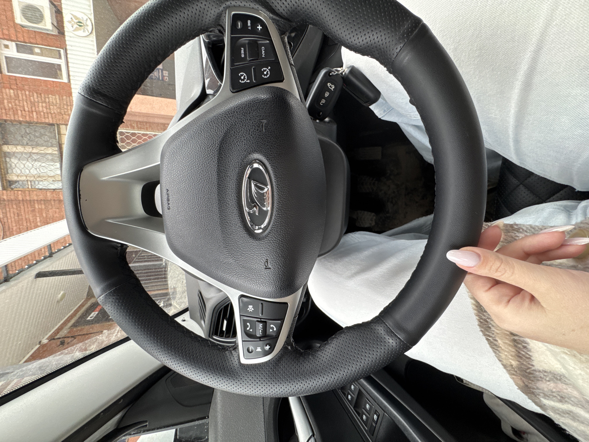 A high-angle, slightly distorted, close-up photograph shows the interior of a vehicle, specifically the steering wheel, partially obscured by a person's hands and lap. The steering wheel is black leather with a silver central component featuring a vehicle logo, control buttons on both sides, and an airbag label. The perforated texture of the leather is visible. A car key is inserted into the ignition. The person's hands are visible on the right side of the frame, with light-colored manicured nails. The person is wearing a light-colored garment. Through the windshield, a blurred view of a brick building is seen, along with what appears to be either window or mirror. Given the car logo, the location could be in Votkinsk, Russia.