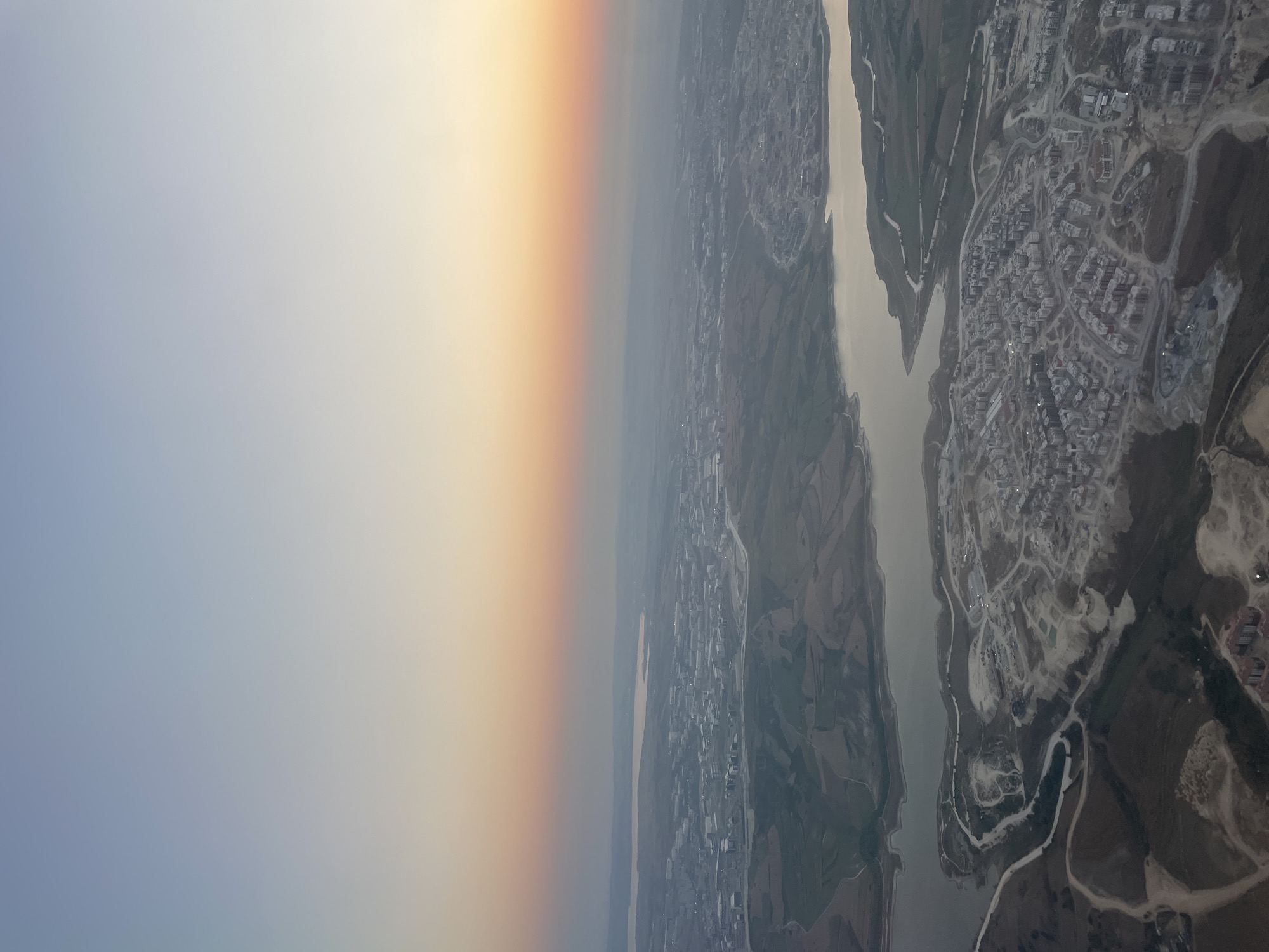 This aerial view depicts a developing urban landscape on the outskirts of Beykoz, Turkey, near a body of water. The scene is bathed in the soft, gradient light of either sunrise or sunset, with the sky transitioning from a pale blue at the top to hues of orange and pink closer to the horizon.
In the foreground, a densely populated residential area is under construction. Numerous apartment buildings, mostly gray and white, are visible, many appearing to be in various stages of completion. The surrounding terrain shows evidence of significant earthmoving and construction activity, with piles of dirt and sand, as well as unpaved roads and pathways snaking through the development.
A large, somewhat murky body of water, likely a reservoir or a section of the sea, lies to the north of the construction zone. Its banks are irregular, with some areas appearing eroded or natural, while others show signs of human alteration.
Further in the distance, more of the city of Beykoz stretches across the landscape, with buildings appearing smaller and less distinct due to atmospheric haze. Green fields and undulated terrain separate different parts of the urban sprawl and the natural landscape. The overall impression is one of rapid development and expansion in a scenic, coastal environment.