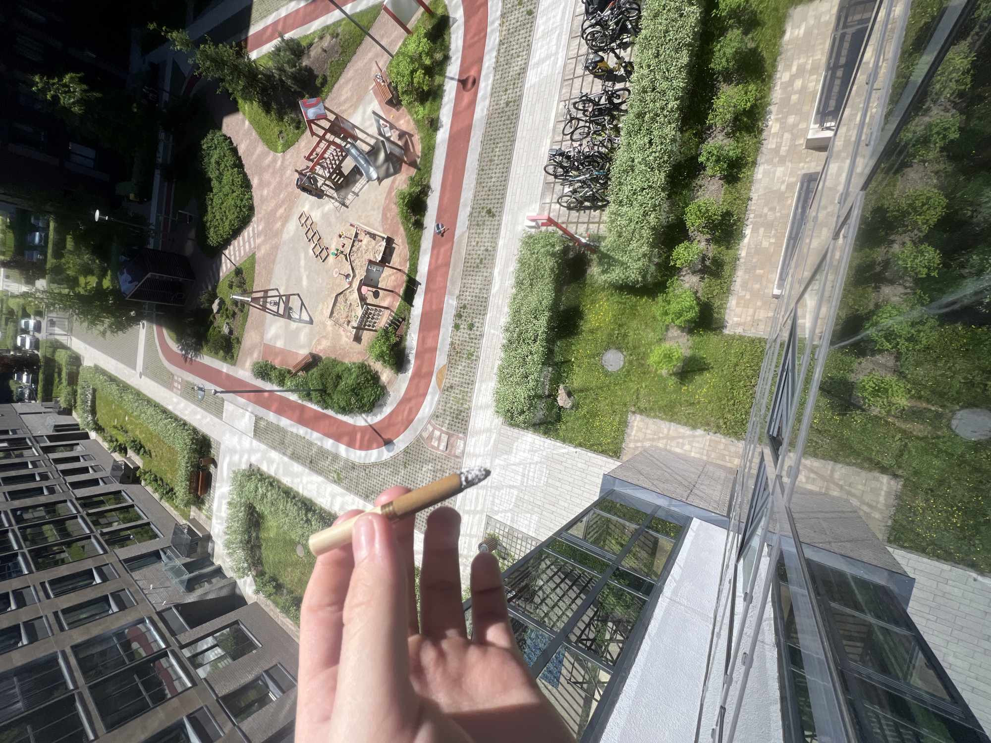 The image provides an elevated, top-down perspective of a modern residential complex and its landscaped courtyard, consistent with contemporary urban developments found in Saint Petersburg, Russia.
In the foreground, a light-skinned hand, likely belonging to an adult, is visible holding a lit cigarette or blunt with a light-colored filter and brown wrapper, exhibiting a visible ash tip. The lower portion of the frame shows reflections from a white wall and glass panels, indicating the photograph was taken from inside a building, possibly through a window or from a balcony with a glass railing.
The main subject of the background is a well-maintained courtyard surrounded by multi-story residential buildings with dark facades and numerous reflective windows. The courtyard features a network of light gray paved walkways and a distinctive red-colored track, likely designed for jogging or cycling, winding through green spaces. These green areas consist of manicured grass, dense hedges, and various trees and shrubs, some with small yellow flowers.
A prominent children's playground occupies a central section of the courtyard, equipped with a sandpit containing small toys, multiple climbing structures, and slides made of metal and wood, some painted red. Other amenities include wooden benches strategically placed along pathways, lampposts, and a dark-roofed covered pavilion or seating area. Near the bottom right, several bicycle racks are densely packed with numerous bicycles. In the farther background, additional residential buildings and a street with parked cars are visible, extending the urban landscape. The scene is illuminated by bright daylight, casting clear shadows.