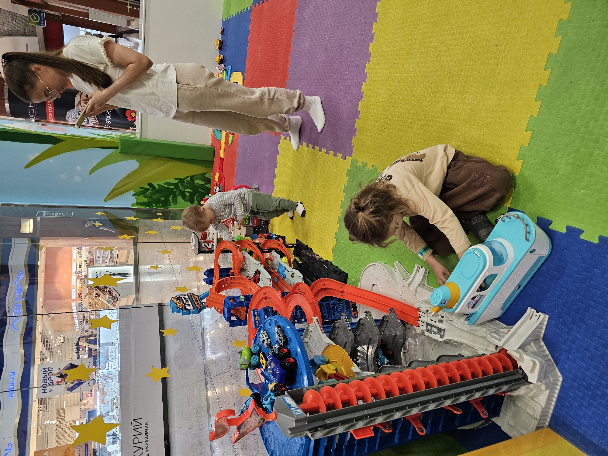 The scene is an indoor play area, possibly in a shopping mall, in Khimki, Russia. There is a woman standing, looking at her phone, and two children playing with a large Hot Wheels track. The track has multiple loops, twists, and turns. The children are positioned on colorful puzzle-piece floor mats. There are yellow star decorations on the glass. The background displays store advertisements.