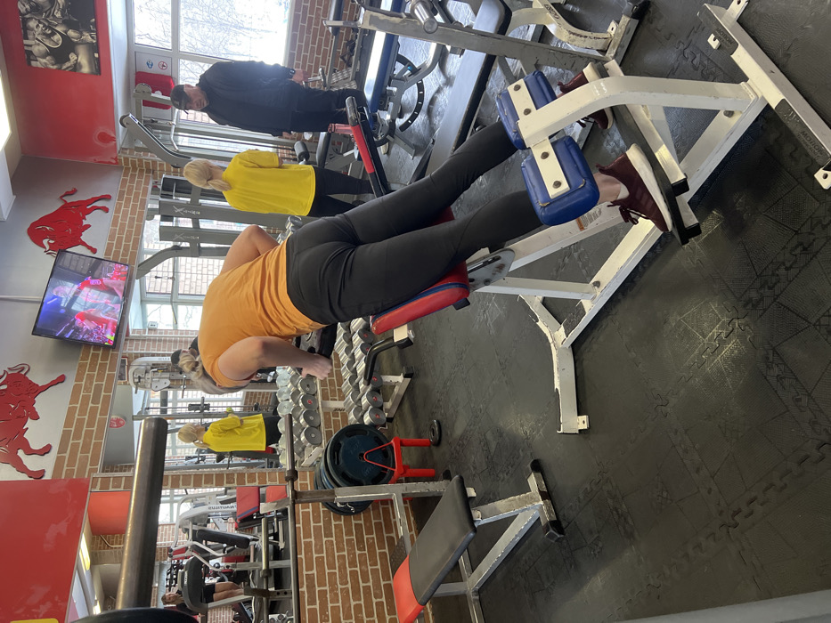The scene is an indoor gym, consistent with facilities found in cities like Saint Petersburg, Russia. In the immediate foreground, a person with light-colored hair, clad in an orange t-shirt and black athletic leggings, performs a back extension exercise on a hyperextension machine. The machine features a white frame with red and blue padding. The individual's back is oriented towards the viewer, revealing maroon athletic shoes.
To the left, a silver utility bench with a black and red upholstered pad rests on the black interlocking rubber mat flooring. Behind the exercising individual, a silver dumbbell rack holds multiple dumbbells, and various barbell plates, including a blue one, are visible near a red stand.
In the midground-right, a woman with light-colored hair, wearing a yellow t-shirt and dark pants, stands opposite a man. The man is dressed in a dark jacket, dark pants, and a dark baseball cap. The two individuals are engaged in an interaction. Large windows on the right wall allow natural light into the space, revealing an exterior consistent with a winter or overcast day.
The gym walls feature sections of exposed brickwork and solid red paint, adorned with red bull silhouette decorations. A flat-screen television, displaying a program, is mounted on the wall. A large mirror on the left wall reflects another gym patron, a woman in dark attire, operating a cardio machine.