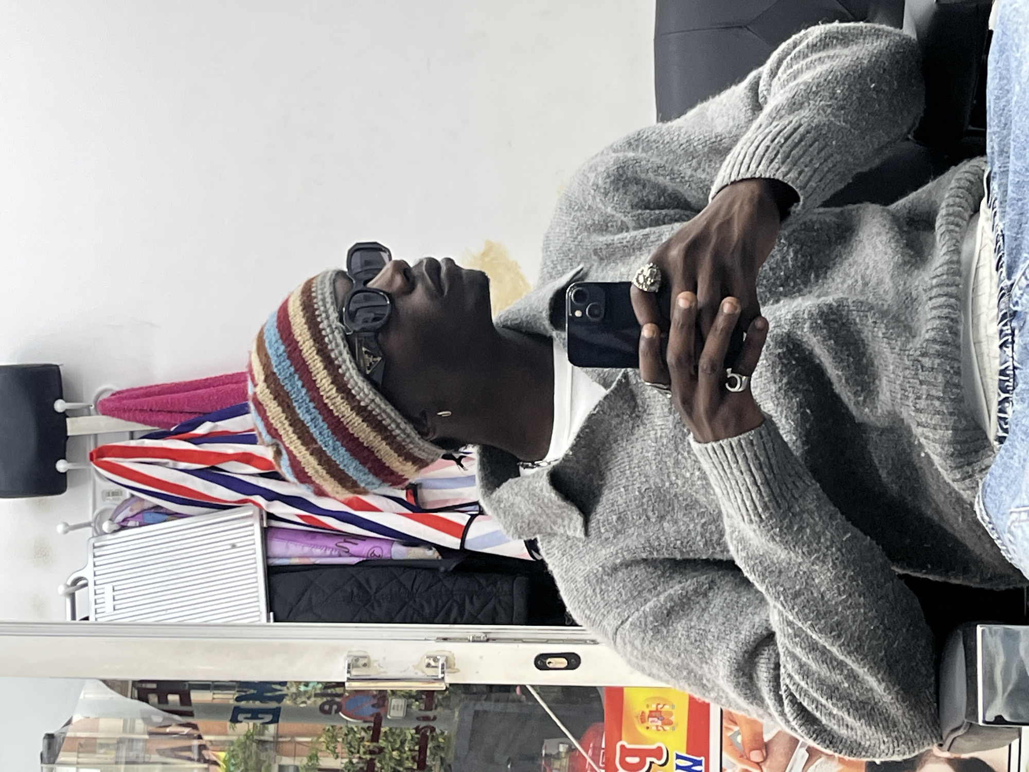 A dark-skinned person wearing a multicolored striped beanie, sunglasses, and a gray sweater is sitting in what appears to be a salon or barbershop. They are holding a black smartphone in their hands, possibly taking a selfie. The person is wearing several rings on their fingers.
Behind the person, various items are hanging, including a pink towel and an umbrella with red, white, and blue stripes. Below these items is a metal tray with a black quilted bag on top.
The location of the image is in Barcelona, Spain. This is evidenced by a window that reflects the street and signage including: a red sign with the letter "B" and "Б", and other letters in Cyrillic script, as well as a faded sign with the Spanish flag.