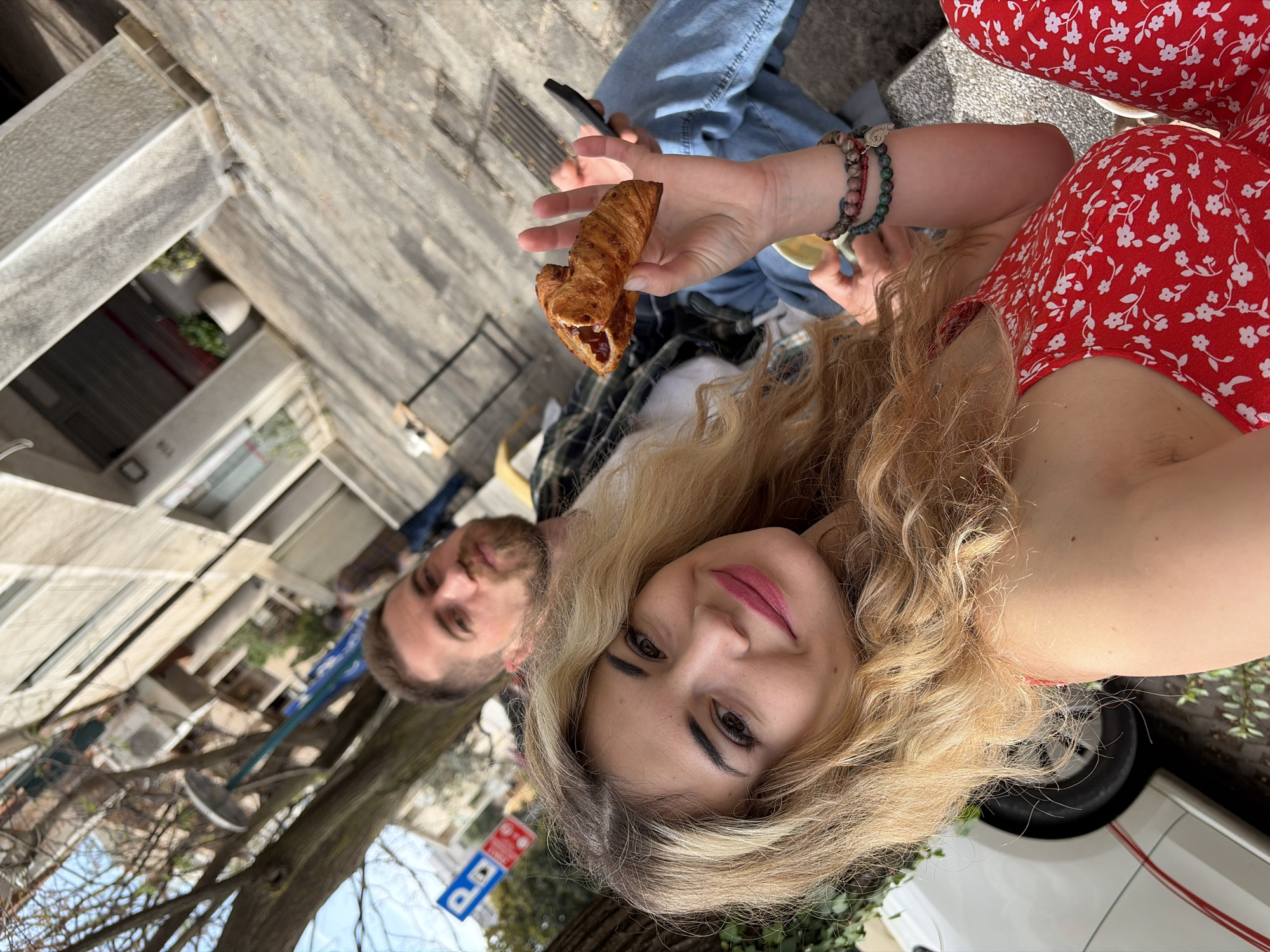 Captured in the city of Budva, Montenegro, the image presents a young couple outdoors, engaging in an activity likely involving food and photography. The female subject, with blonde hair and wearing a red floral dress, is holding a pastry, possibly a croissant, filled with jam in one hand. In the background, a fair-skinned male with a beard is visible. The couple appears to be seated, possibly on a curb or low wall, evidenced by the jeans and casual wear of an unseen person. A smartphone is in the background. The backdrop reveals an urban environment with buildings, trees, and street signs, typical of a European city setting.