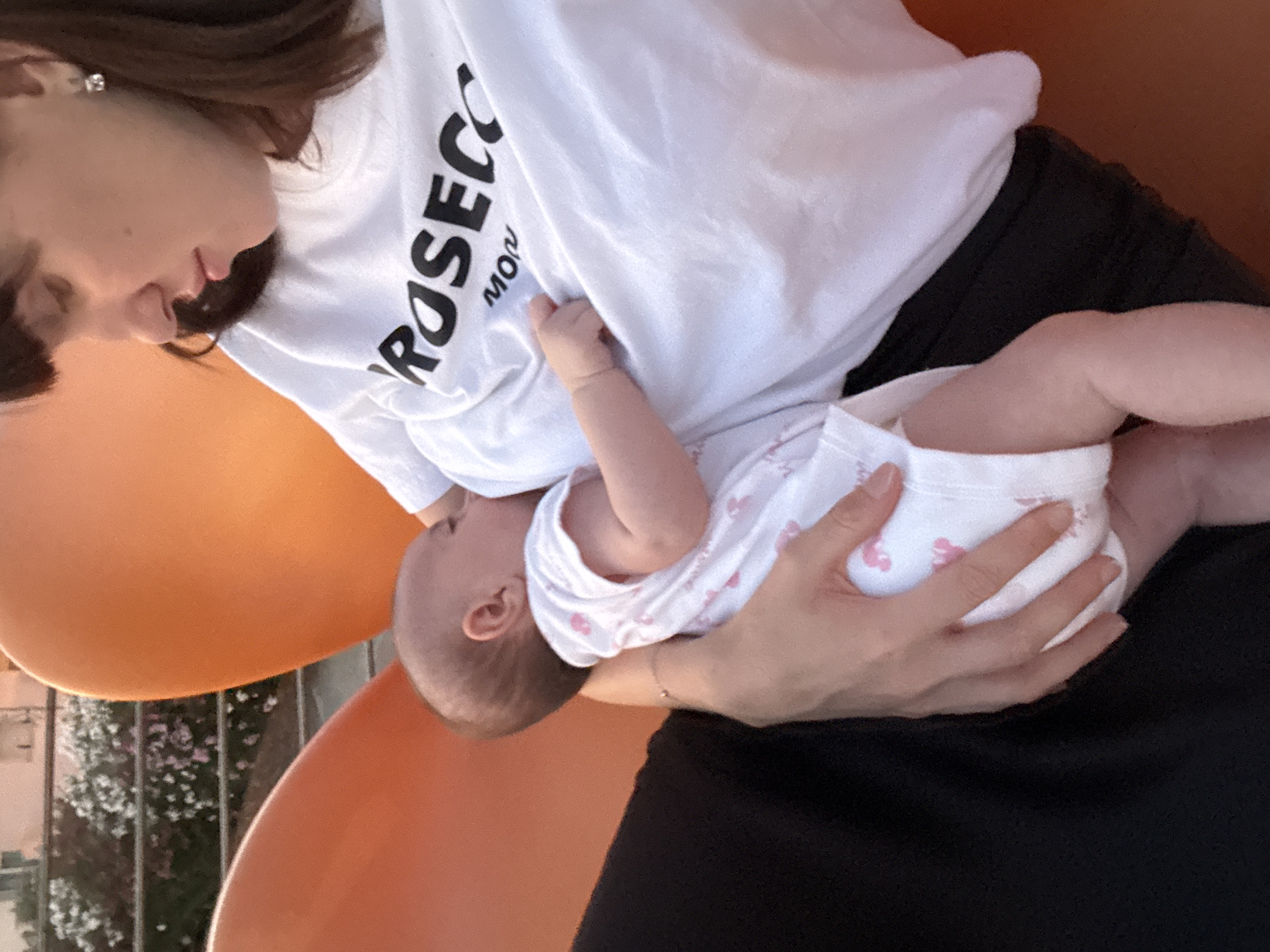 The image shows a woman breastfeeding a baby. The woman is wearing a white t-shirt with the word "PROSECCO" printed on it in black lettering. The baby is wearing a white onesie with pink pattern. They are sitting on an orange chair outdoors, with a railing and some flowering plants in the background. The location is Isola delle Femmine, Italy. The woman's arm is supporting the baby, and her hand is visible on the baby's back. The baby's head is turned towards the woman's chest. The overall lighting suggests it is daytime, possibly late afternoon or early evening, given the warm tones.