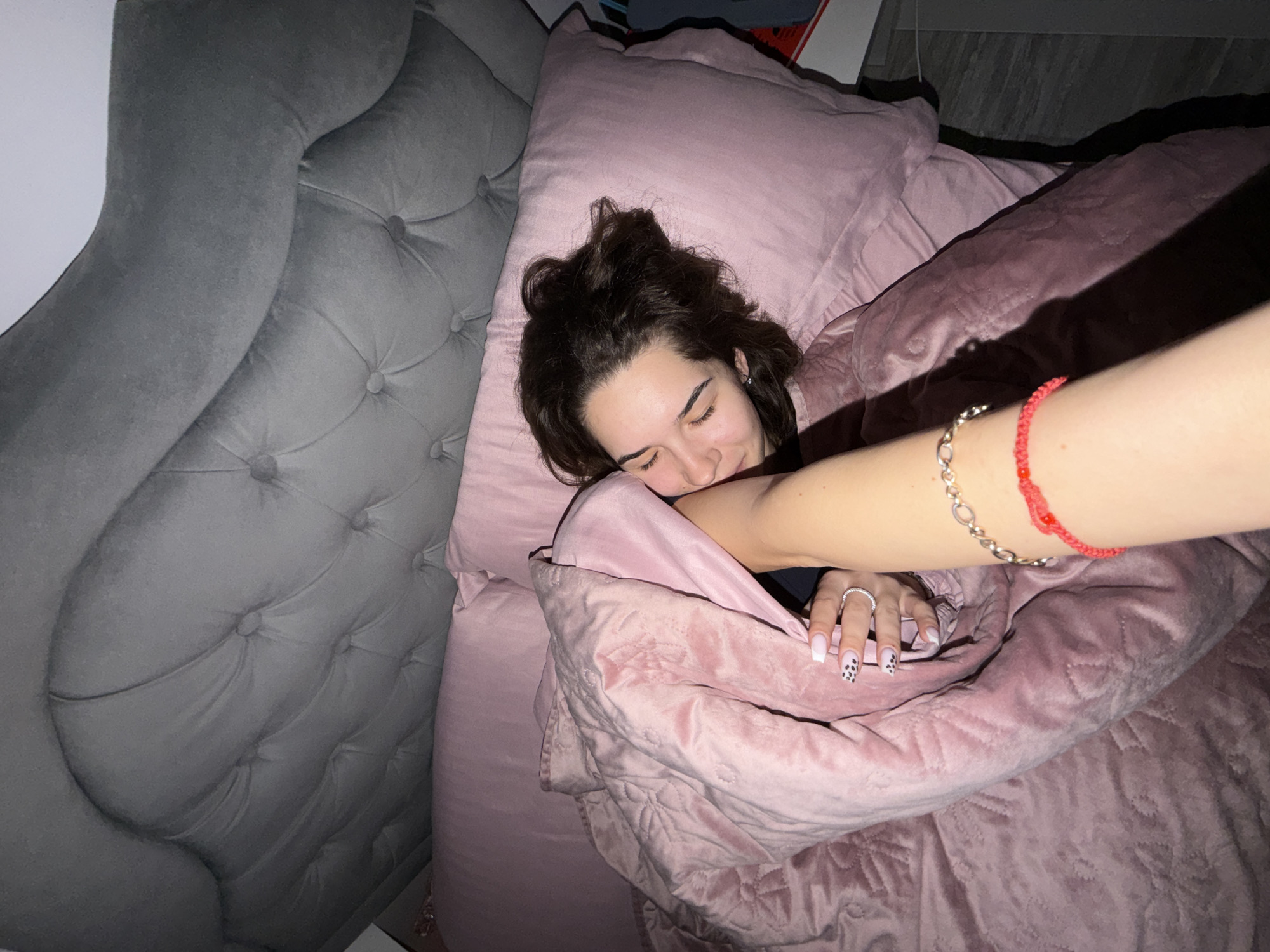 A high-angle shot shows a woman lying in a pink bed with a gray tufted headboard. She is partially covered by a pink quilt. One arm, adorned with bracelets and a ring, extends across the quilt towards the camera. The image's location is unverifiable.
