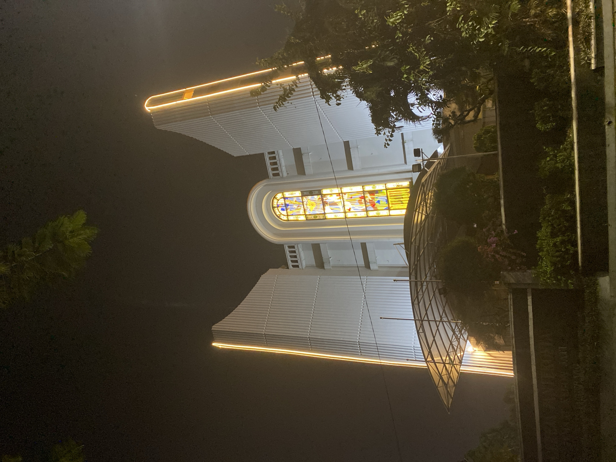 This is a night-time, low-angle, exterior shot of the Church of Christ the King (Gereja Kristus Raja) located on Jalan Maulana Hasanuddin in Tangerang, Banten, Indonesia. The facade of the church is illuminated by bright, vertical orange lights on its tall, white, ribbed side structures. A large, arched stained-glass window, glowing with yellow and multi-colored light, is visible in the central upper section of the building. In front of the church, a translucent, sloped roof structure is supported by metal poles. The foreground consists of a dark wall, greenery, and part of a street. The sky is dark and overcast.