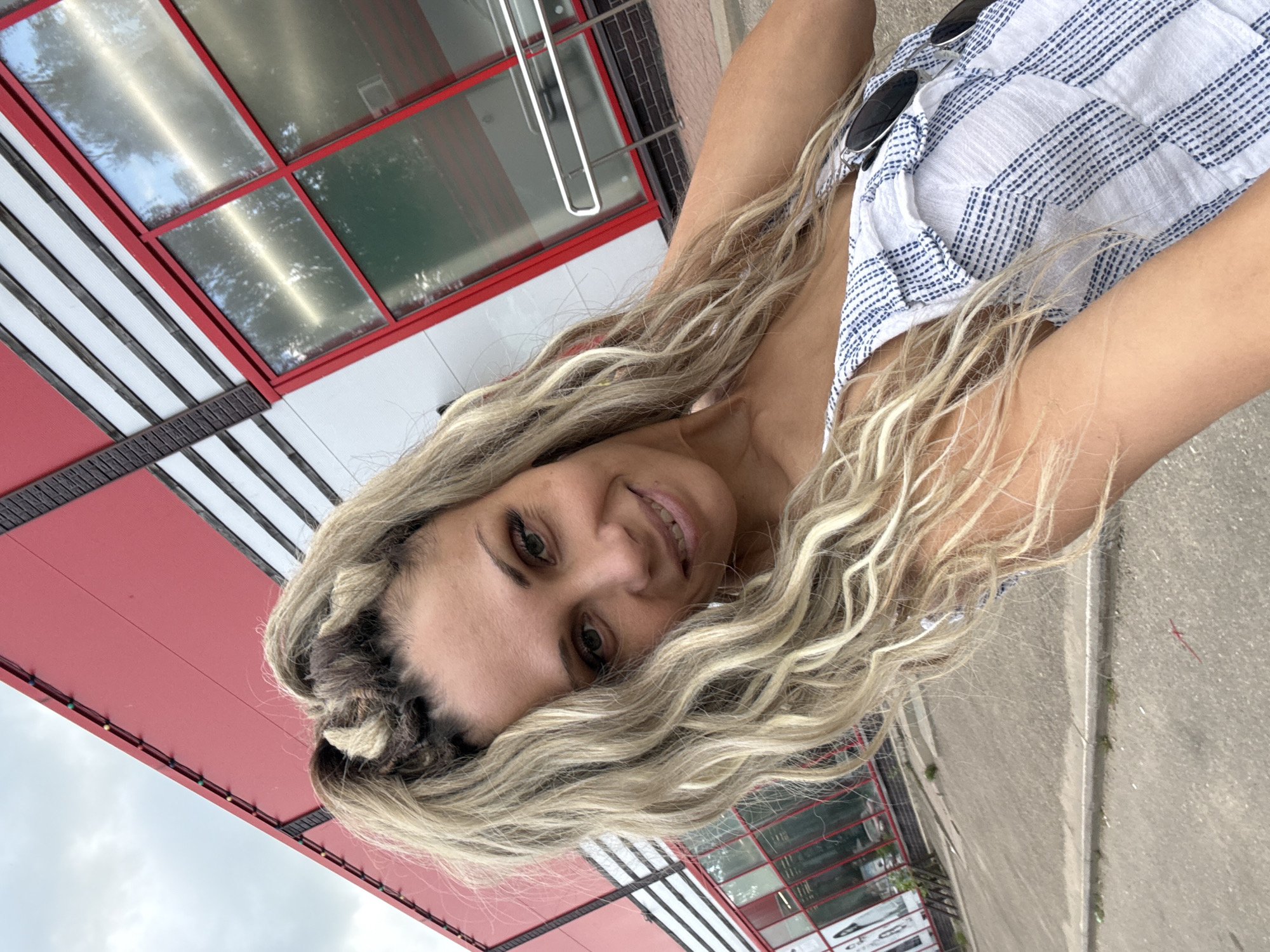 A blonde woman with braided hair and curly strands poses for a selfie outdoors in front of a building with a red facade and large windows. She is wearing a blue and white striped top and sunglasses are tucked into the neckline. The ground is paved, and there is a sidewalk or curb visible. The overall environment suggests an urban or commercial setting.