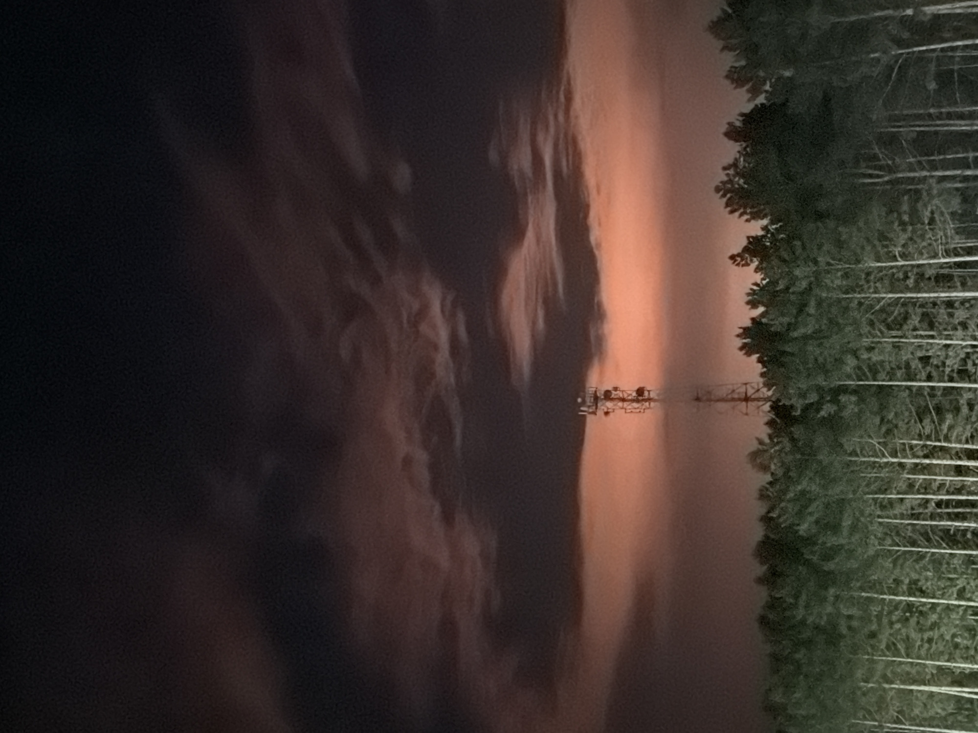 An outdoor, long shot photograph captures a telecommunications tower silhouetted against a dramatic sky. The tower stands tall and skeletal, composed of a lattice of metal beams, with multiple platforms and antennas protruding from its structure. It is positioned above a dense line of dark green pine trees that form a horizontal band across the lower third of the image. The sky above the trees is a striking gradient of deep dark blue at the top, transitioning into lighter shades of gray and pinkish-orange as it approaches the horizon. Wispy, dark clouds are scattered across the sky, some catching the light and appearing to glow with an orange hue. The overall impression is one of a remote, possibly rural, landscape at dusk or dawn, with the man-made structure of the tower contrasting with the natural elements of the forest and sky. The scene appears to be located in the vicinity of Tsiolkovsky, Russia, given the provided context.
