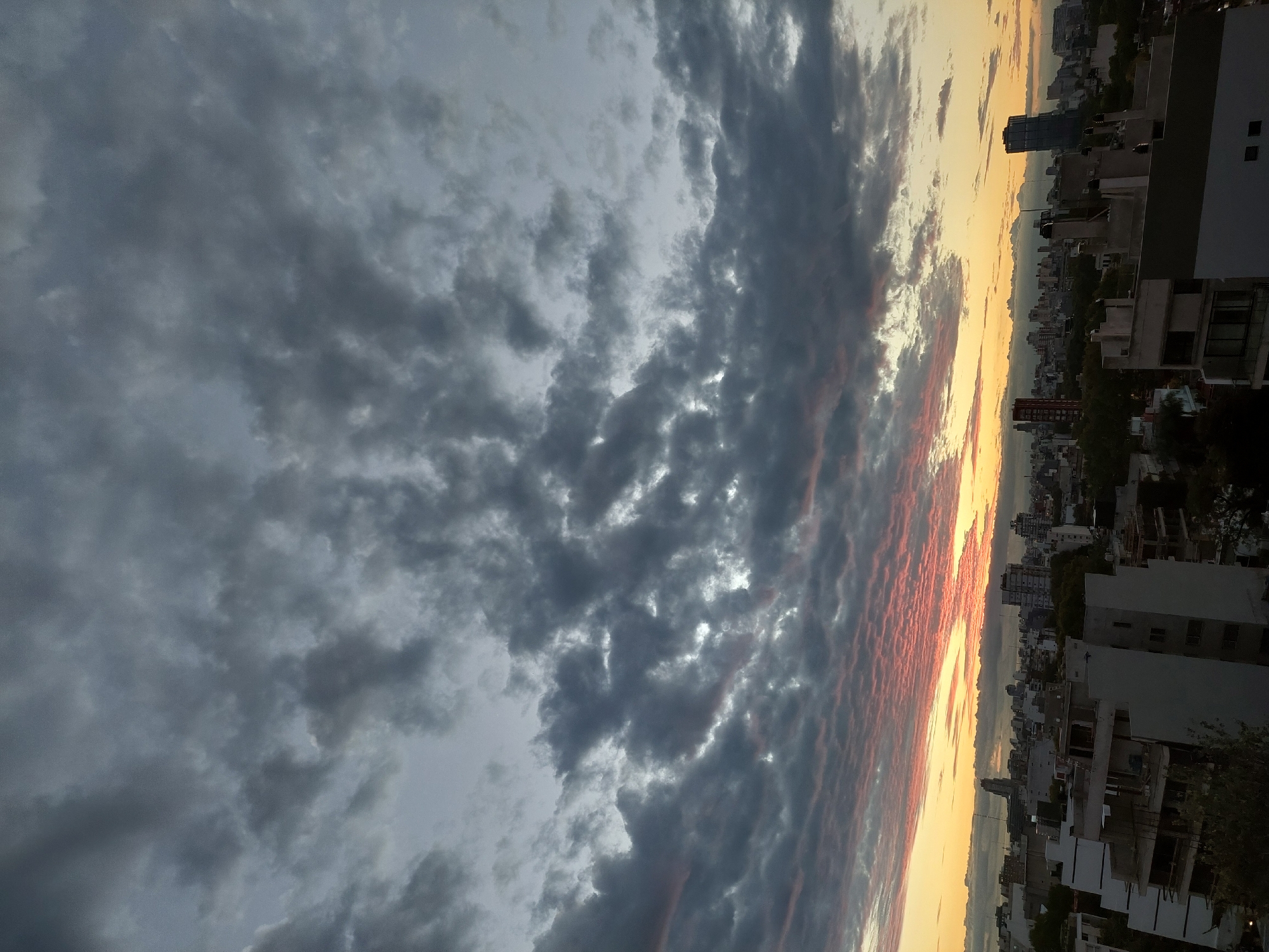 The image captures an urban skyline during a dramatic sunset, likely in Buenos Aires, Argentina. The foreground is dominated by a sky filled with dark, textured cumuliform clouds on the left, transitioning into a vibrant sunset on the right. The sunset displays hues of yellow, orange, and streaks of red.
Below the horizon line is a cityscape composed of buildings and structures of varying heights and architectural styles. Trees are visible in between the buildings. The shot appears to be taken from an elevated position, providing a panoramic view of the city.
No people are clearly visible in the image.