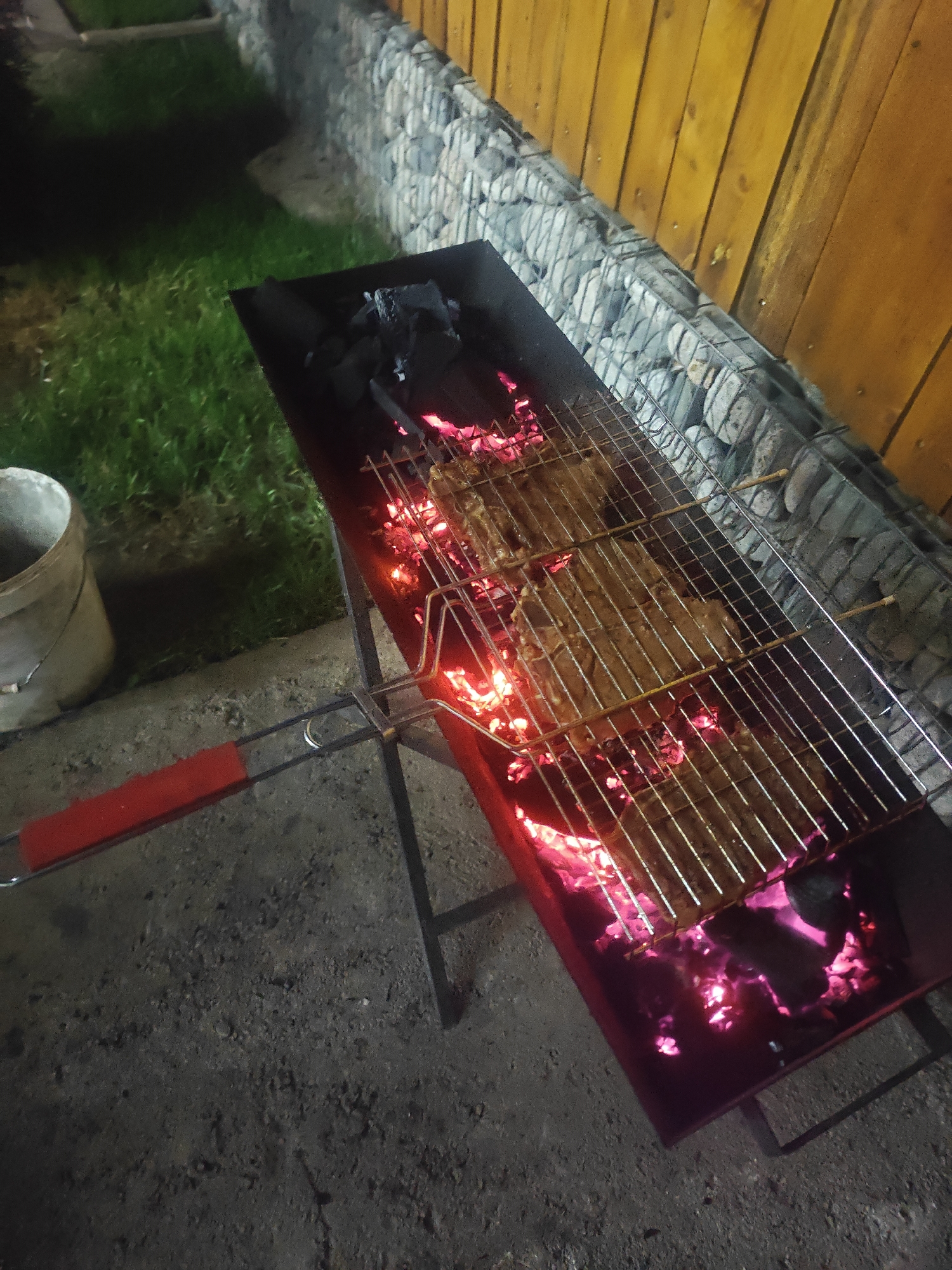 An overhead, high-angle shot shows a long, rectangular barbecue grill with a metal grate holding several pieces of raw meat. The grill is filled with glowing red coals, creating a vibrant pink and red hue beneath the meat. A red-handled tool, possibly a barbecue fork, is positioned to the left of the grill, extending towards the viewer. In the background, a gabion wall constructed with wire cages filled with stones separates the grilling area from a grassy lawn on the left and a wooden fence on the right. To the far left of the frame, a portion of a light-colored bucket is visible. The scene appears to be outdoors, at night, with artificial lighting illuminating the area. The ground beneath the grill and extending to the left is a rough, textured surface, possibly concrete or dirt. The overall impression is of an outdoor cooking setup in a backyard or patio area, possibly in a residential setting. The specific location of Байгелди, Kyrgyzstan, is not directly evident from the visual cues within the image itself.