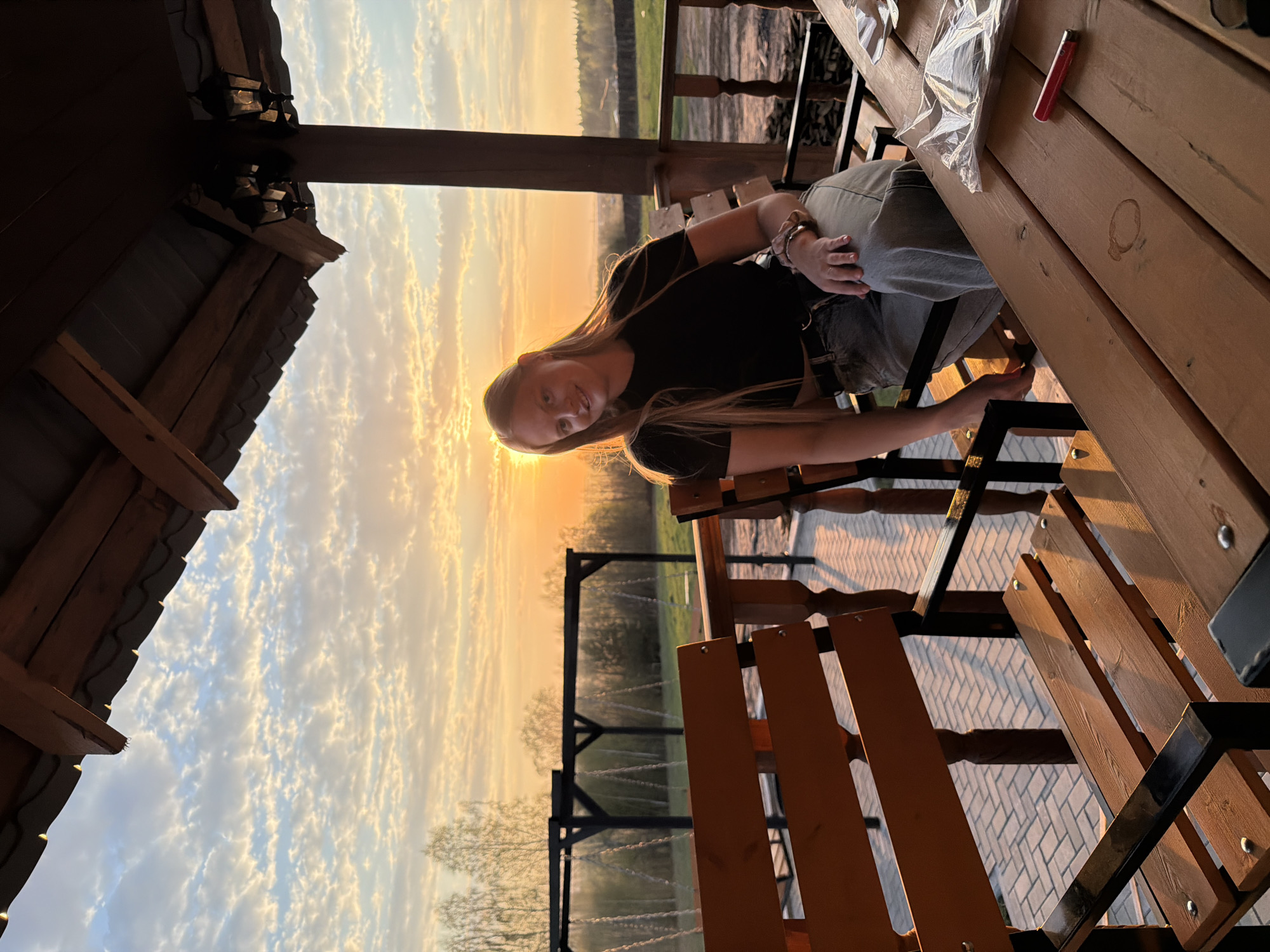 A young woman with long blonde hair sits at a wooden picnic table within an open gazebo in Kamennoye, Russia. The gazebo has a sloped, dark roof. The woman is wearing a black t-shirt and light-colored jeans. She's facing the camera, with a slight smile. Outside the gazebo, the scene is bathed in the light of a sunset or sunrise. The sky is filled with streaks of light and clouds. A swing set is visible in the background. The ground is paved with light-colored bricks. A red lighter and a plastic bag are on the table next to her.