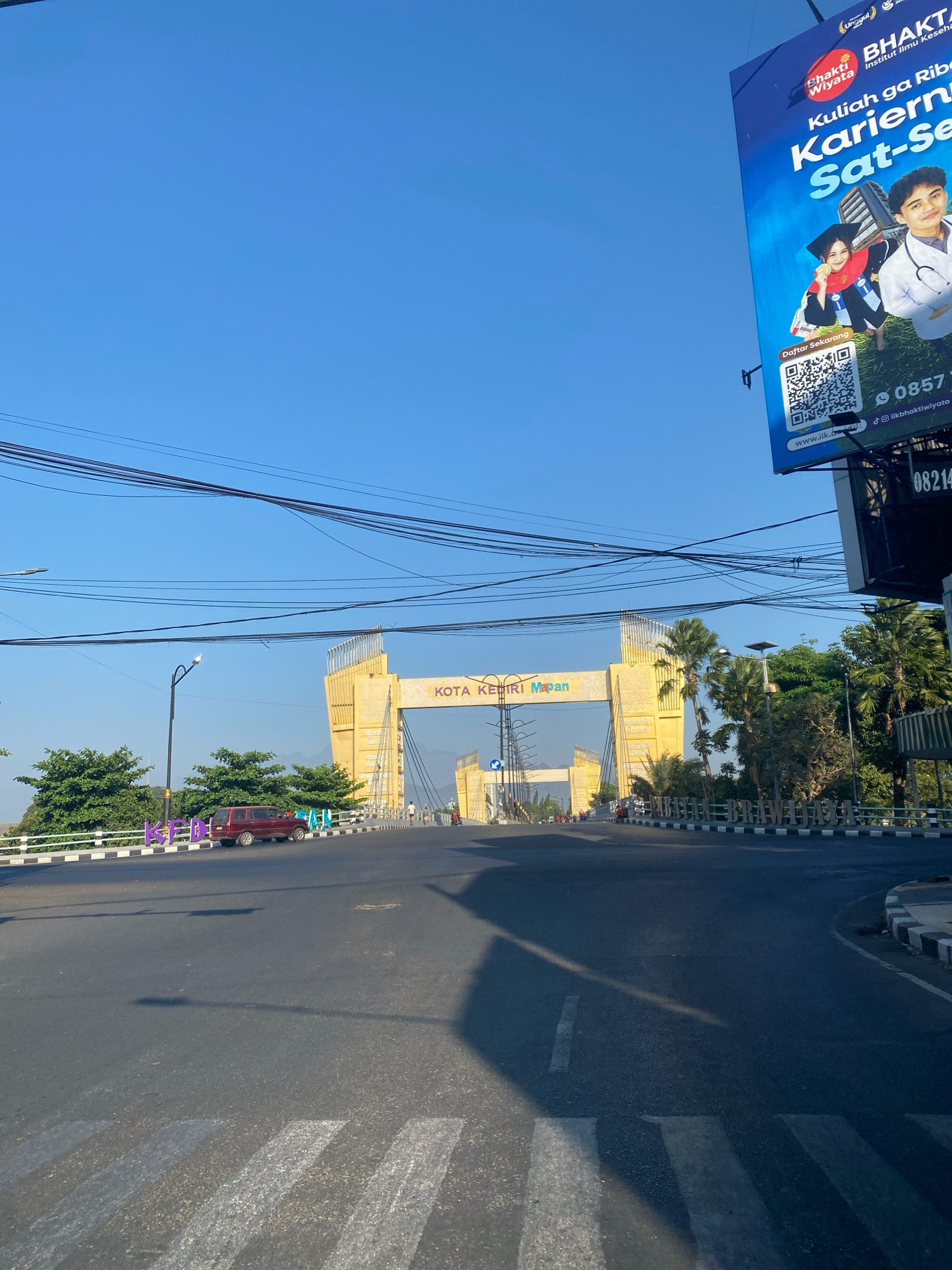 The image shows a street scene in Kediri City, Indonesia. In the background, a large yellow archway spans the road with the words "KOTA KEDIRI Maju" visible on it. To the right of the archway, a billboard for "Bhakti Wiyata" displays advertisements for educational programs and contact information. The foreground shows a road with white crosswalk markings and a red SUV parked on the side. Trees and power lines are visible throughout the scene, under a clear blue sky.