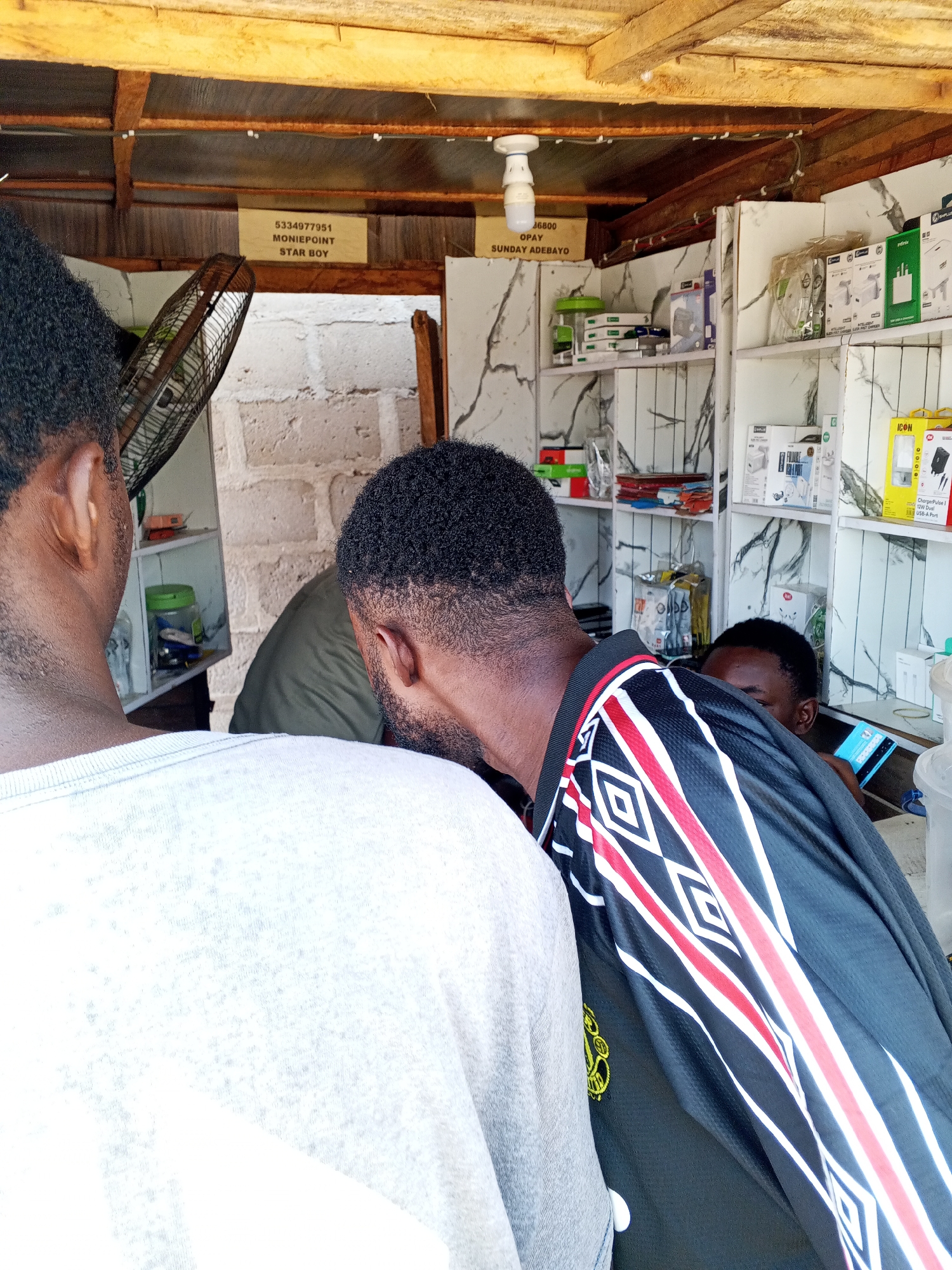 The image depicts the interior of a small retail space, likely a phone or electronics accessory shop, in Lokoja, Nigeria. Several people are clustered within the shop, partially obscuring the view. From left to right, a man's head and shoulder are visible, wearing a light gray shirt. Next to him, a man is visible wearing an olive green t-shirt. A third man stands in the foreground, wearing a black shirt with a pattern of white and red diamonds along the sleeves. Peeking behind them is another man with a cell phone in his hand. The shelves are filled with various packaged items, including chargers and other electronic accessories. Two signs are visible above; one reads "5334977951 MONIEPOINT STAR BOY" and the other reads "46800 OPAY SUNDAY ADEBAYO". A fan is mounted to the wall on the left, and a light bulb hangs from the wooden ceiling. The walls behind the shelves are white with a marble-like pattern.