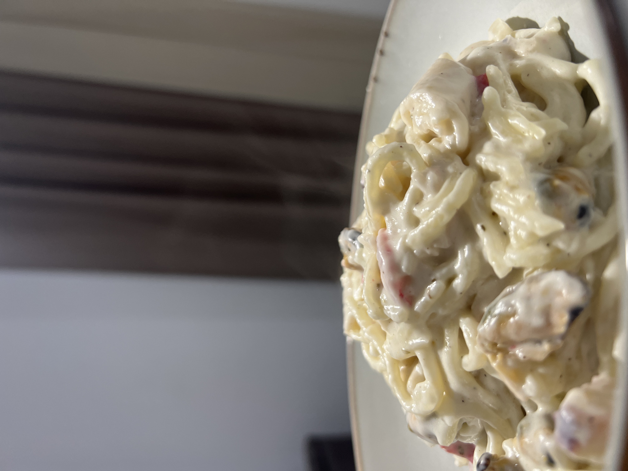 The image presents a close-up, slightly elevated view of a prepared pasta dish served in a light-colored bowl within an indoor environment. The pasta, appearing as spaghetti or linguine, is generously coated in a thick, off-white, creamy sauce. Interspersed throughout the pasta are several mussels, some partially visible within their dark shells, along with small, diced red pieces consistent with bell pepper. Faint wisps of steam are discernible rising from the dish, indicating it is hot. The bowl has a light-toned interior and a slightly darker, textured or dotted rim. The background is out of focus, consisting of a plain light grey wall on the left and dark brown pleated curtains on the right. No individuals are present. The precise location within Budva, Montenegro, cannot be determined from the visual evidence.