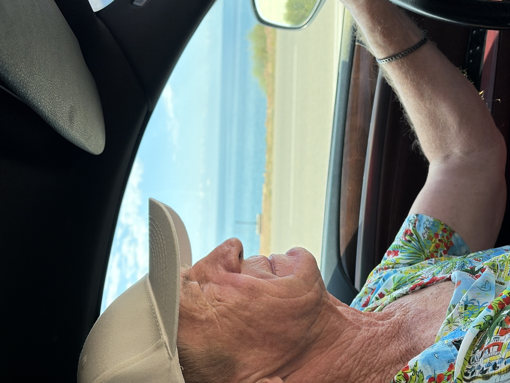 A medium shot captures an elderly Caucasian man driving a car. He is wearing a light-colored baseball cap and a brightly colored shirt with a tropical print. The man's profile is visible as he looks out the window. The car's interior is visible, with a wooden trim on the dashboard and a side-view mirror. Outside the car, a body of water is visible, likely the ocean or a large lake, with a clear blue sky above. The scene appears to be set in a coastal area, possibly on a road trip.