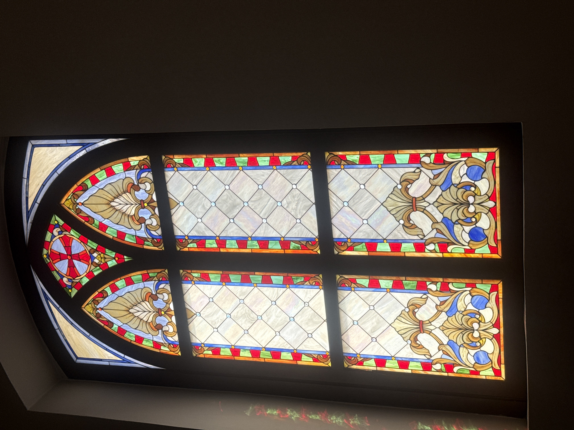 A detailed view of a stained-glass window with six panes. The panes are arranged in two rows. The first row consists of a decorative half-oval pane on the left, followed by two rectangular panes. The bottom row mirrors this arrangement. The half-oval panes feature a cross on the left and stylized plant designs, while the rectangular panes contain a diamond pattern. The environment is dark and unclear. The location is Unknown, Unknown.