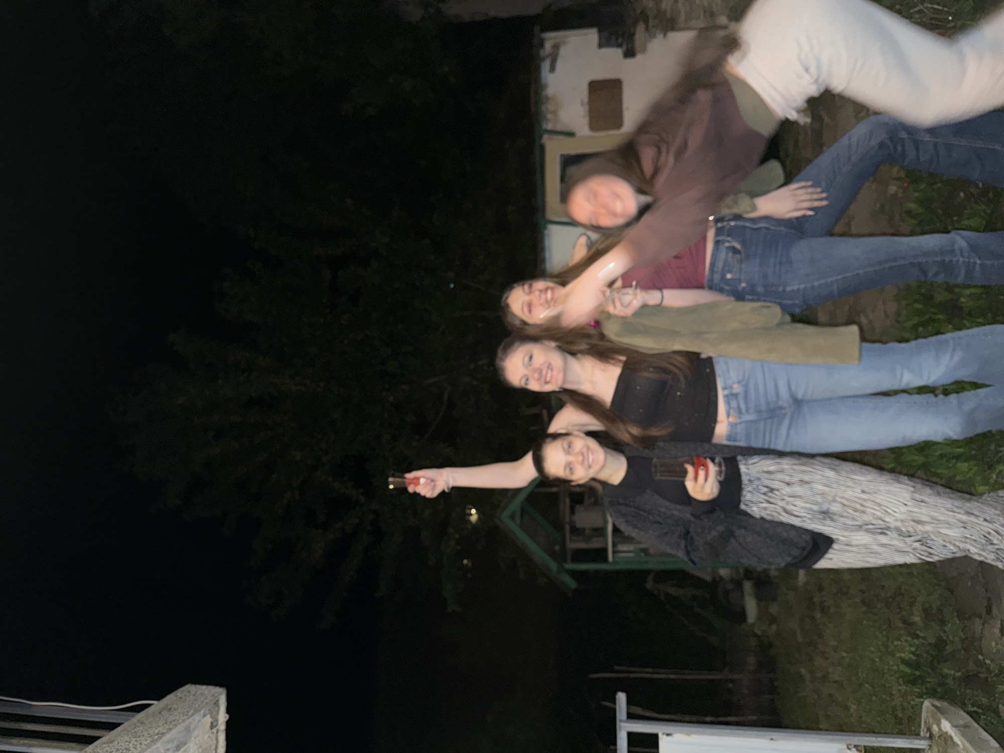 The image captures four young adult females outdoors at night in Tsar Kaloyan, Bulgaria. The background is dark, featuring silhouettes of trees and indistinct foliage, along with two small structures: a green wooden shed or playhouse visible to the left of the center, and a lighter-colored building with a dark opening further back on the right. A white vertical post or structure is partially visible on the far left.
From left to right:
1. A female, smiling, wears a dark top, a dark patterned open cardigan, and light-colored, vertical-striped, wide-leg trousers. She holds a short-stemmed glass containing a red liquid in her right hand.
2. A female, smiling, wears a black strapless top and blue flared jeans. Her right arm is raised, holding a champagne flute with a red liquid.
3. A female, smiling with an open mouth, wears a dark red top and blue flared jeans. An olive-green jacket is draped over her left shoulder and arm. She appears to be holding an object with her right hand, which is extended towards the person on the far right.
4. A female is in motion, captured with significant blur, leaning forward and smiling. She wears a dark brown long-sleeved top over a lighter undershirt and light-colored trousers. Her right arm is extended towards the third female, suggesting an interaction or gesture.
All subjects appear to be in good spirits, smiling or laughing, indicative of a social event or gathering. The scene is illuminated by an unseen artificial light source, highlighting the individuals against the dark nocturnal environment.