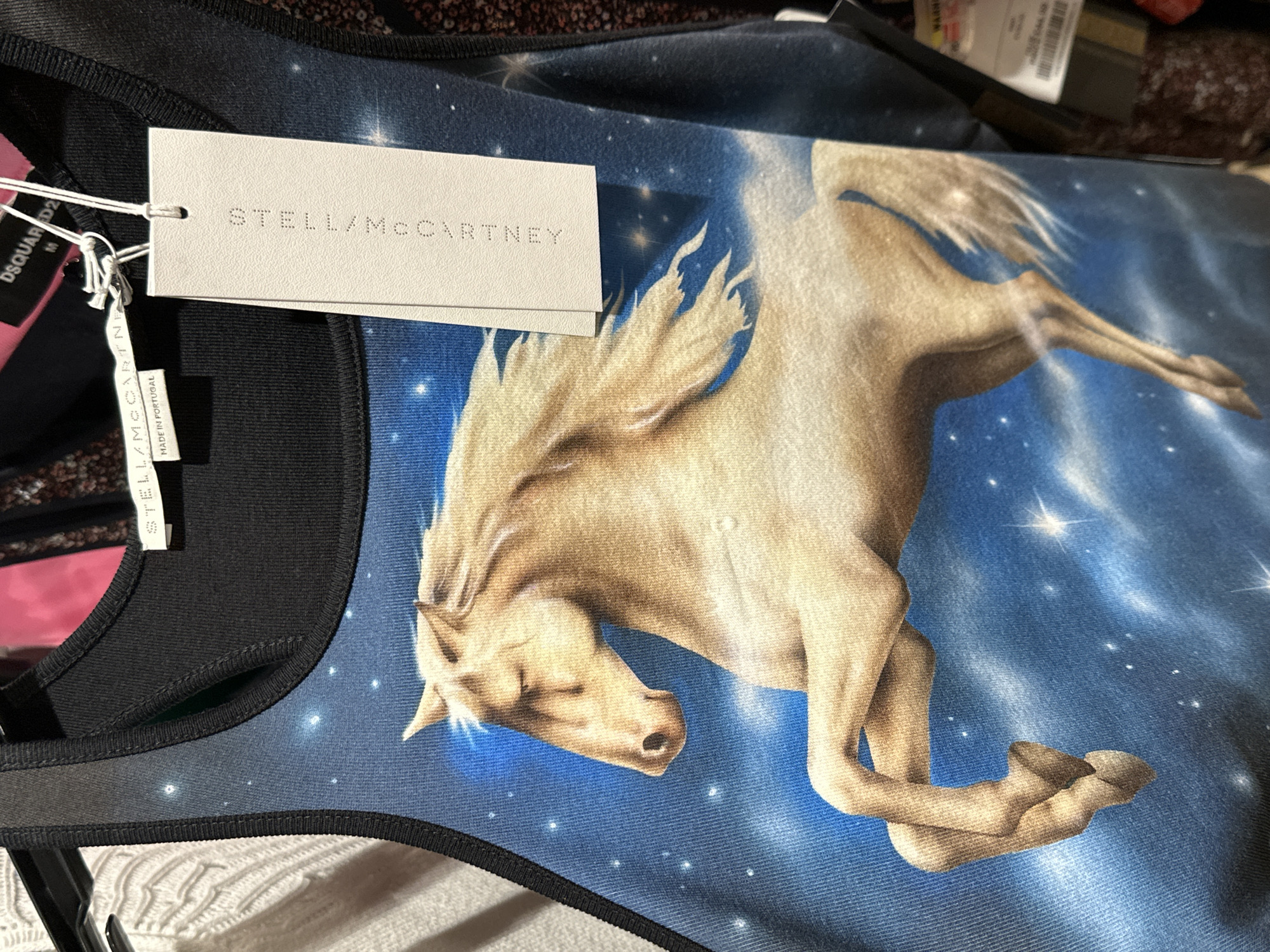 The image shows a close-up of a Stella McCartney garment, likely a top, featuring a print of a flying horse (Pegasus) against a starry, blue sky background. The garment has black trim. A Stella McCartney tag is attached, along with a DSQUARED2 tag. The garment appears to be hanging on a rack, with other garments visible in the background. The environment is likely a retail setting. No people are visible in the frame. The specific location in London, United Kingdom, cannot be determined from this image.