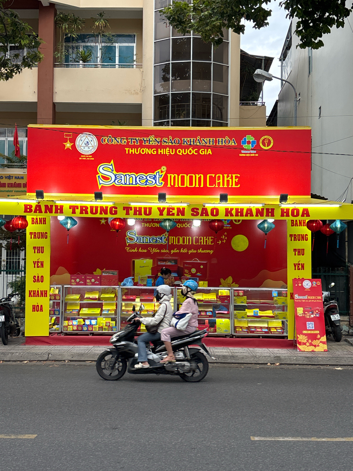 In Nha Trang, Vietnam, on a cloudy day, a street-level view captures a store selling Sanest Moon Cakes. The storefront is adorned with a large red banner featuring the brand name "Sanest MOON CAKE" in prominent yellow lettering, along with Vietnamese text. The establishment displays rows of moon cakes in colorful boxes, arranged on shelves behind glass. Decorative red and blue lanterns hang from the awning, and a bright yellow vertical sign on the left lists "BANH TRUNG THU YEN SAO KHANH HOA," indicating the product and origin. Two individuals on a black motorcycle are captured in motion in the foreground, passing in front of the store. The motorcycle rider wears a white helmet and jacket, while the passenger is dressed in a pink jacket and white helmet. A second motorcycle is parked to the right of the store. The background shows a multi-story building with a balcony and trees.