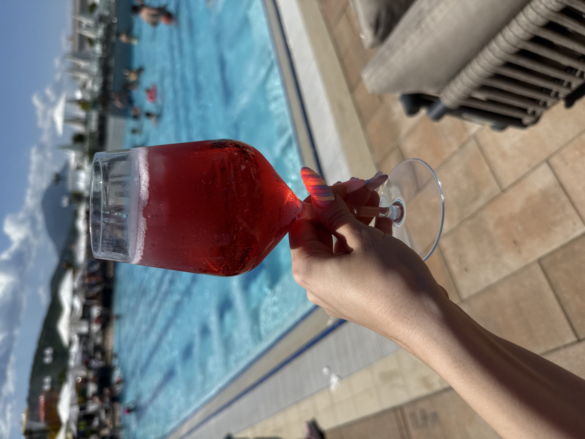 A person's right hand, with multi-colored manicured fingernails, is centrally positioned in the lower frame, holding a stemmed glass. The glass contains a translucent red beverage with ice cubes and a white foamy head, consistent with a cocktail. The hand and glass are in sharp focus. The background is out of focus, depicting a vibrant blue swimming pool with multiple individuals swimming and relaxing. Beyond the pool, white parasols and indistinct buildings suggest a resort environment. In the far background, hazy mountains are visible under a blue sky with scattered white clouds. The scene is illuminated by bright daylight. To the right of the hand, a portion of a wicker-style lounge chair with light-colored cushions is visible, out of focus. The poolside ground is covered with light brown square tiles.