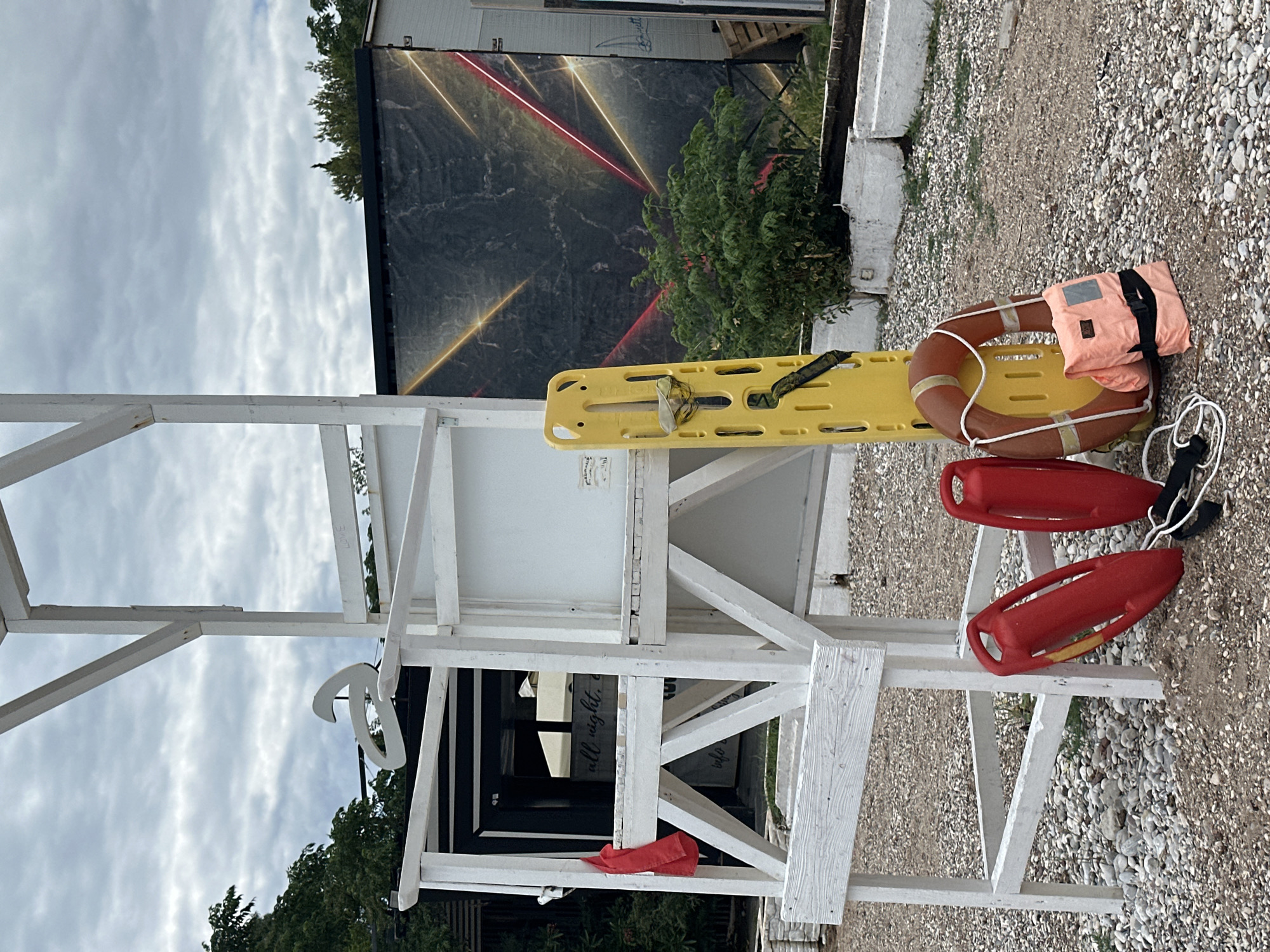 The image, taken in Budva, Montenegro, displays a white wooden lifeguard-style stand or observation post under an overcast sky. The multi-tiered structure, built from painted wooden beams, occupies the center-foreground.
Various pieces of rescue equipment are arranged on and around the stand. On its mid-level platform, a bright yellow backboard with straps is positioned. Adjacent to it, a brown lifebuoy (life ring) with a white rope holds a coral-colored life vest, neatly folded with black straps. Near the base of the stand, two red rescue tubes (torpedo buoys) with attached ropes rest on a lower beam. A small red cloth is also visible tucked into the lower frame. A decorative white curvilinear object hangs from an upper beam.
The ground surrounding the stand is covered in light-colored pebbles or gravel. In the background, behind the stand, a large dark banner or wall features abstract geometric patterns of bright red and yellow/gold light streaks. Green foliage is visible near the base of this banner. Part of a black and white striped structure is faintly visible through the stand's framework.
The scene is set during the daytime, under a cloudy or overcast sky. No people are present, but the arrangement of equipment suggests preparedness for water safety or beach activities. Faintly visible text that appears to read "all right" is present on a white panel behind the yellow backboard.