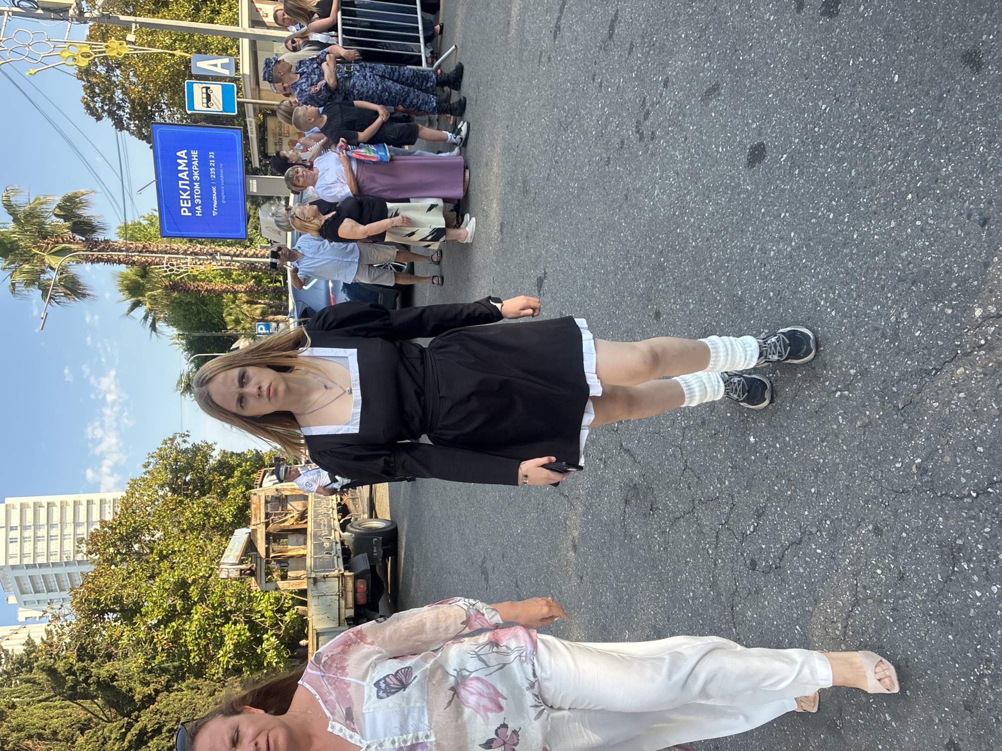 In the city of Sochi, Russia, on a sunlit day, a group of people are gathered on a paved street. In the foreground, a young woman with blonde hair and a serious expression walks towards the camera, wearing a black dress with a white collar and cuffs, white socks, and sneakers. To her left, another woman, partially visible, walks in the same direction, wearing a white patterned blouse and white pants. Behind them, a truck with a crane is parked. Further in the background, a crowd of people, including individuals in uniform, stand near a metal barrier. A large digital billboard displaying advertisements is visible on the right side of the street, with palm trees and a modern building in the distance. The overall scene appears to be an outdoor public event.