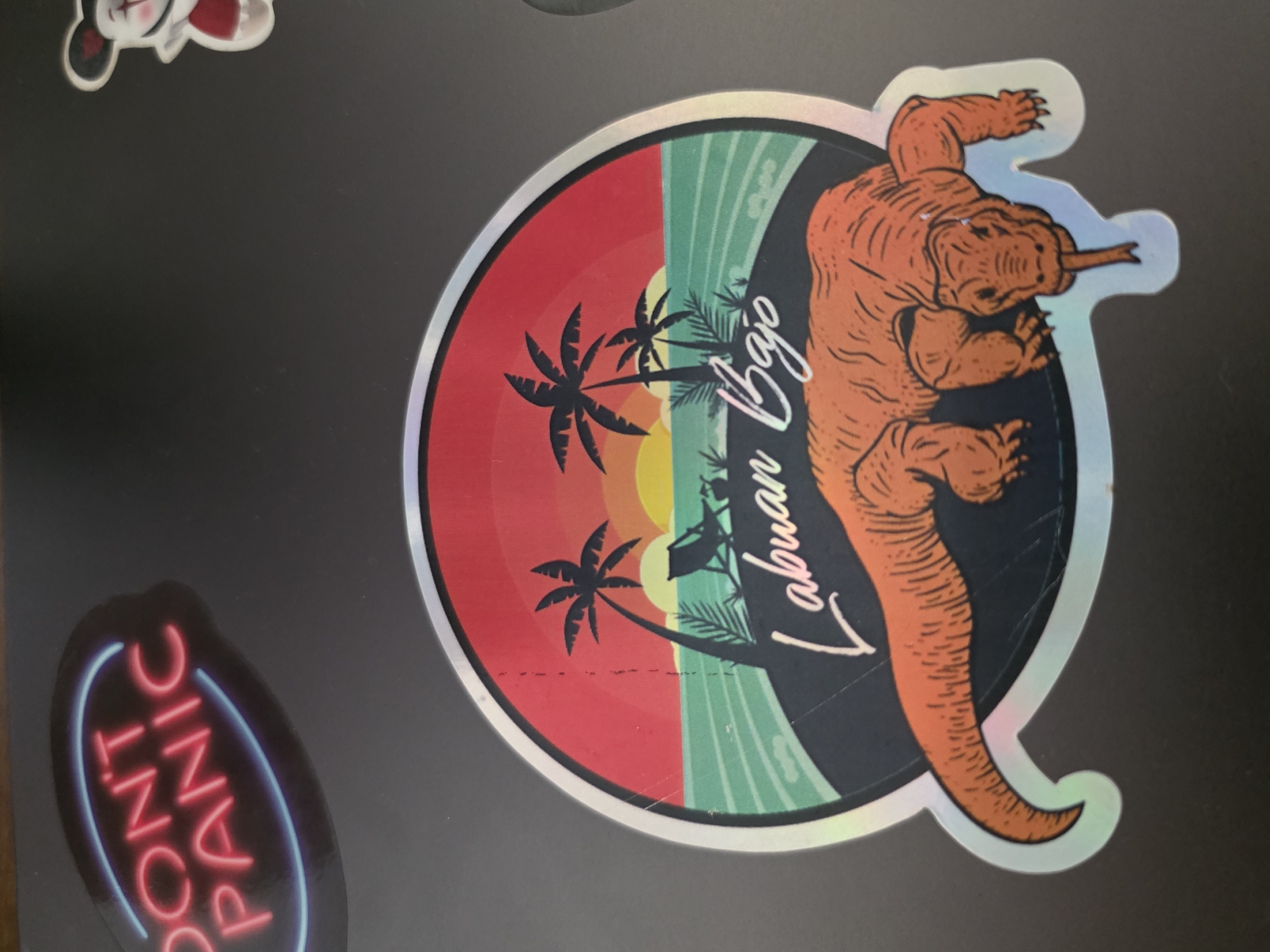 The image shows a laptop with several stickers on it. One sticker is round with a holographic border, depicting a sunset scene with palm trees on the left, a light green wave pattern in the center, and an orange Komodo dragon on the right. The words "Labuan Bajo" are written vertically over the wave and dragon. Another sticker, on the lower left, says "DON'T PANIC" in neon blue and red letters. There's also a partially visible sticker of a cartoon character in the top left corner.