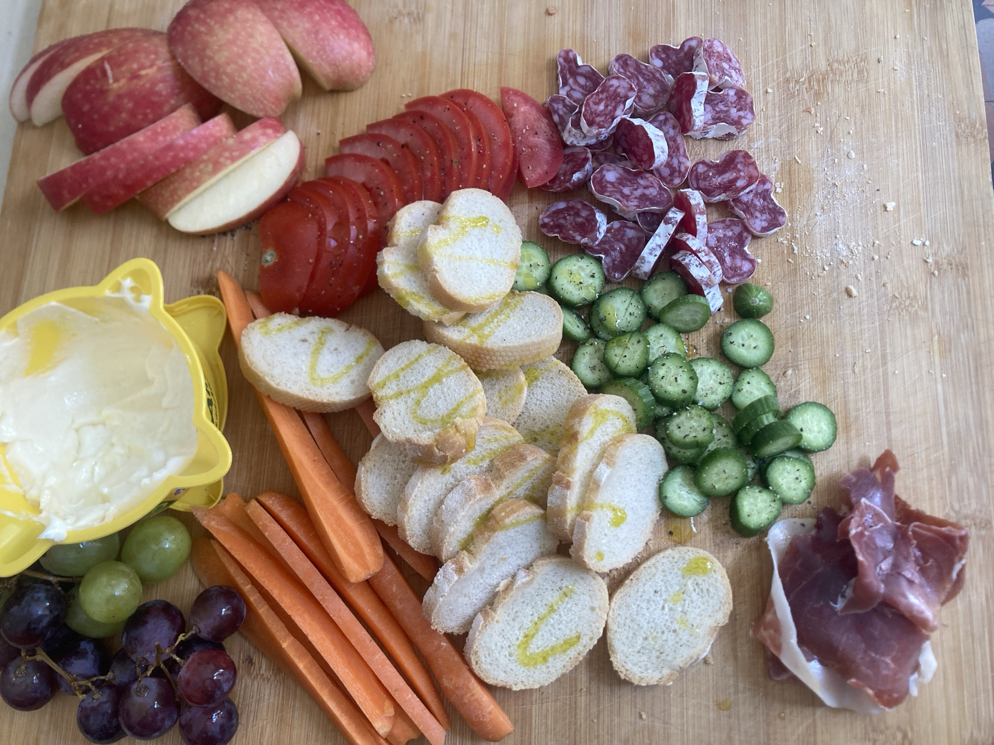 A wooden cutting board is topped with an array of food items. Sliced red apples and tomatoes are positioned at the top left. A yellow bear-shaped container filled with cream cheese is placed adjacent to clusters of green and purple grapes. Carrot sticks lay next to the cream cheese and overlapping bread slices. Sliced cucumbers sit next to the bread slices. A pile of dried sausage slices and ham is at the upper and lower right, respectively. The scene is in a residential setting. The image was taken in Barcelona, Spain. There are no people visible in the image.