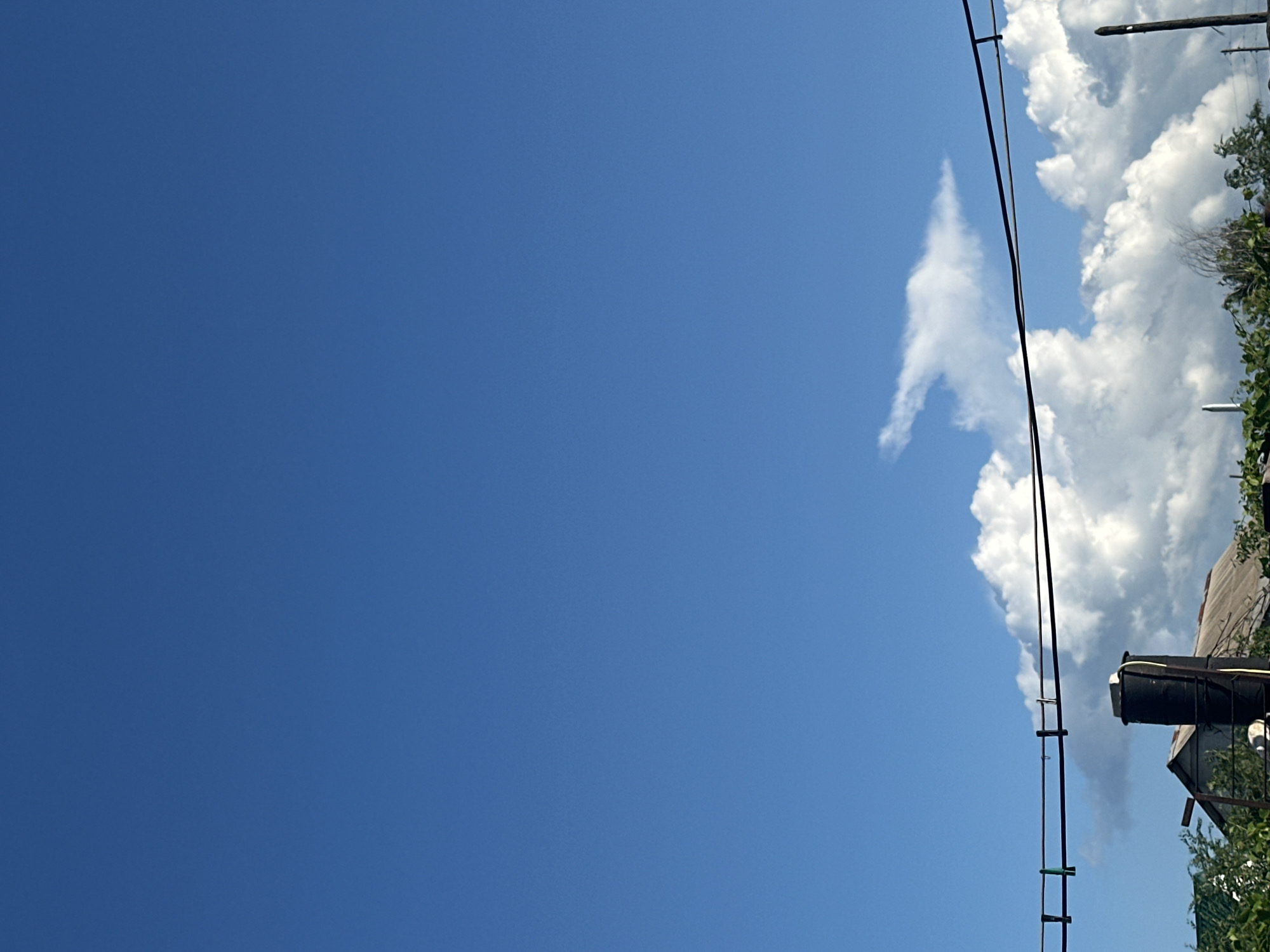 The image depicts a clear, bright blue sky on a sunny day, with a few fluffy white clouds visible on the right side. A power line runs diagonally across the frame from the top right to the bottom left, with a few small clips attached. Below the power line and clouds, there is a glimpse of a building with a grey, weathered roof and some green foliage. A black cylindrical object, possibly a water tank, is mounted on a structure next to the building. The overall scene suggests an outdoor setting, likely in a rural or suburban area given the presence of the power line and the building. The bright sky and distinct clouds indicate it is daytime, likely mid-morning or afternoon. There is no visible text in the image.