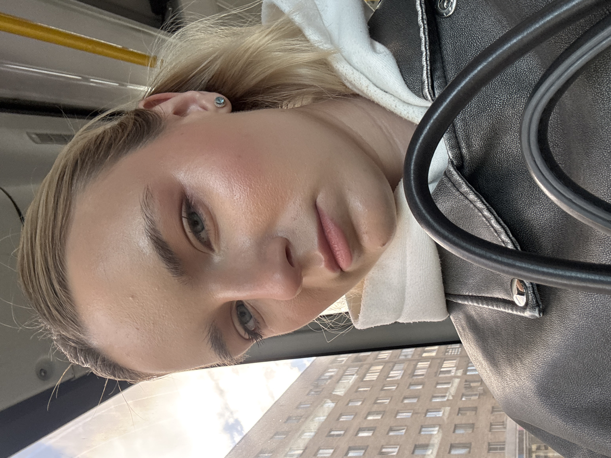A close-up, angled selfie of a fair-skinned woman with blonde hair pulled back, wearing a white hoodie and a black leather jacket. She is looking slightly downward towards the camera. In the background, a tall building with multiple windows is visible through what appears to be a window. A black, circular object, possibly a handle or support, is positioned in the foreground to the right of the woman's face. No information indicates this picture was taken in Vsevolozhsk, Russia.