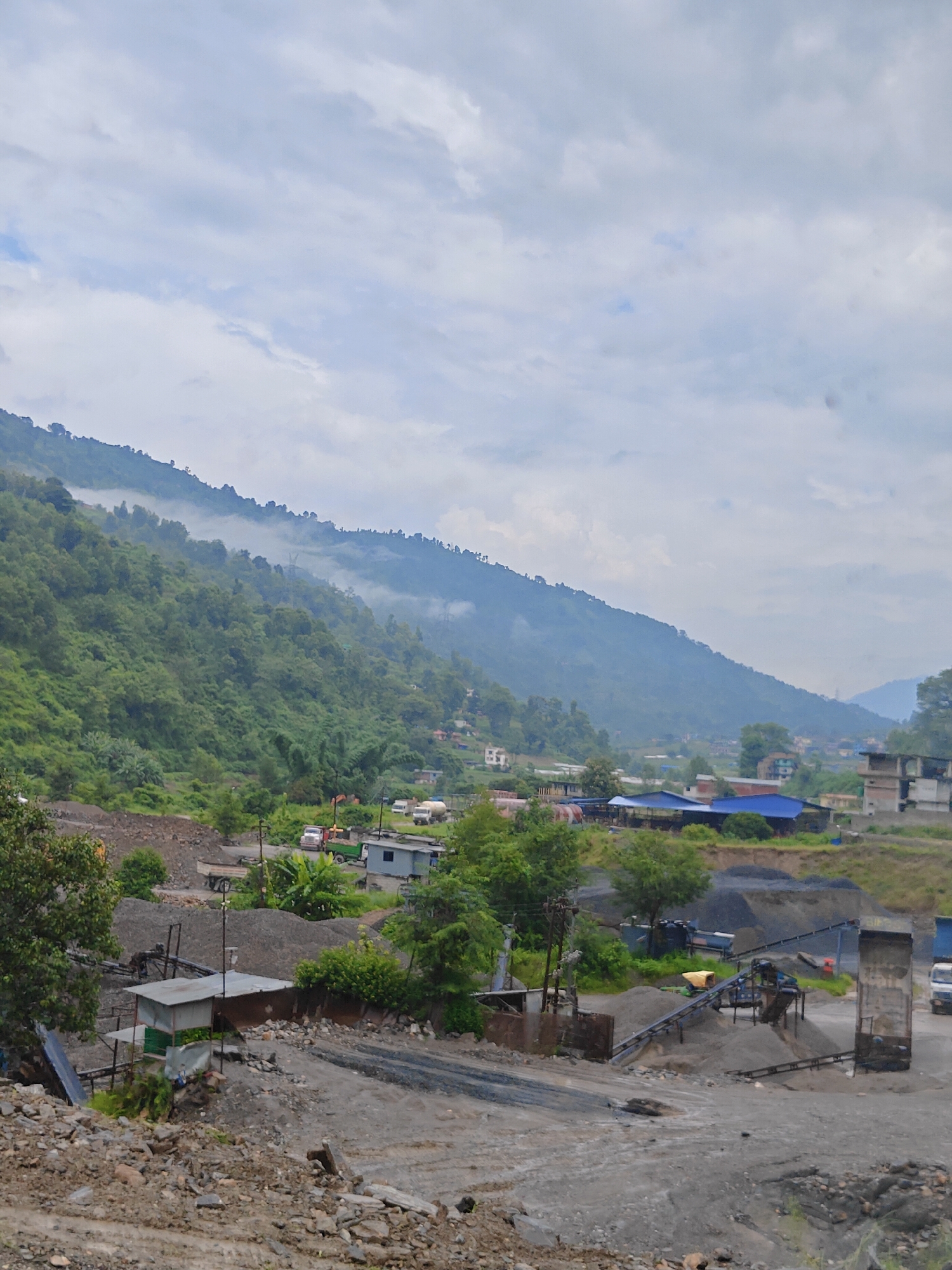The image shows a quarry site in Thakre, Nepal. In the foreground, piles of gravel and rock are visible, along with some industrial equipment, including conveyor belts and crushers. A small shed-like structure is also present. In the middle ground, there are several trucks and a water tanker. Beyond the quarry, a hillside covered in lush green trees rises steeply. Some buildings and houses are scattered on the lower slopes of the hill, with a few visible further up. The sky is overcast with clouds, and a light mist or fog hangs over the higher parts of the mountain. The overall scene depicts an active industrial operation within a natural, mountainous landscape.