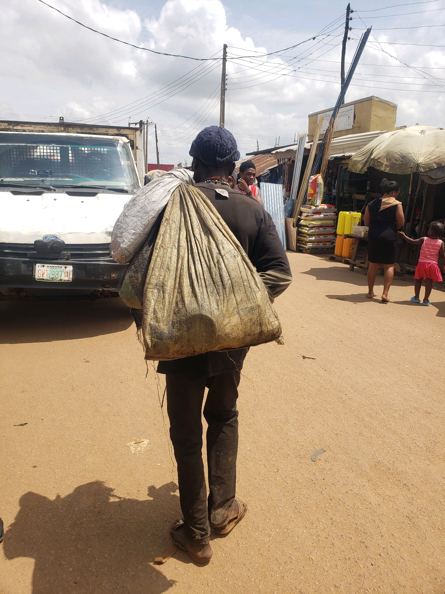 In Sabon Tasha, Nigeria, a person wearing a dark long-sleeved shirt and dark trousers carries two large, heavy bags over their left shoulder. One bag is a lighter colored, possibly woven sack, and the other is a larger, darker, textured sack. The person is walking away from the camera on a dusty, unpaved street. To the left, a white Ford truck is parked, and its license plate reads "KADUNA GRT-987VU". In the background, market stalls with corrugated metal roofs are visible, along with stacks of goods including what appear to be bags of fertilizer or grain and yellow containers. Power lines crisscross the sky, supported by utility poles. Further in the background, a woman in a black dress and a young girl in a pink and white striped top and pink skirt are interacting. The lighting suggests it is daytime, with bright sunlight casting shadows.