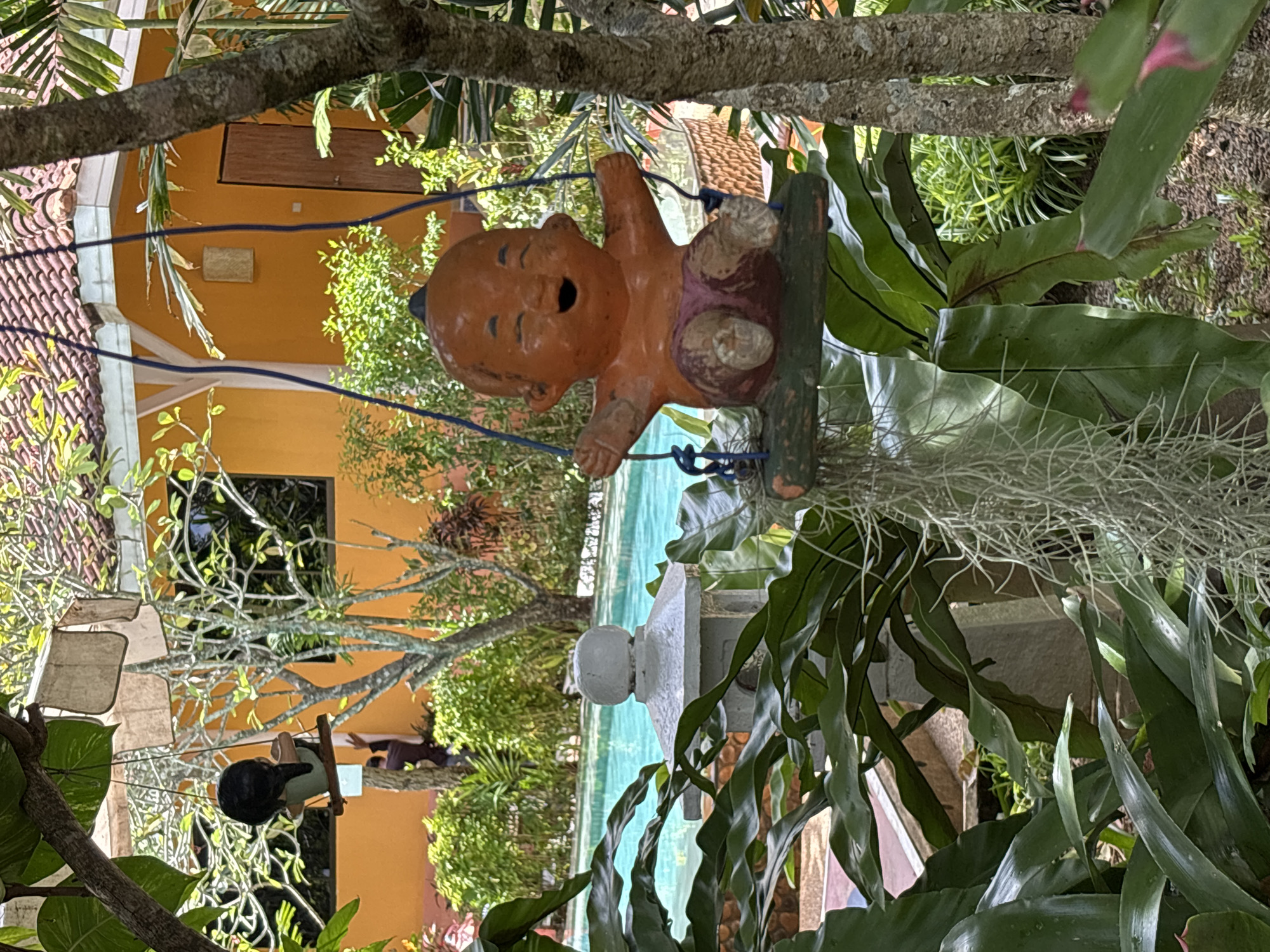 In Chalong, Thailand, a ceramic figurine of a laughing child on a swing is prominently displayed outdoors. The figurine, with its orange skin, open mouth, and closed eyes, is seated on a weathered wooden swing suspended by blue ropes. The swing hangs from a tree branch, partially obscured by lush green foliage. In the background, a swimming pool with turquoise water is visible, adjacent to an orange building with a tiled roof. A white, pagoda-style lantern stands near the pool. The scene is set amidst a tropical garden with various plants and trees, bathed in natural sunlight.