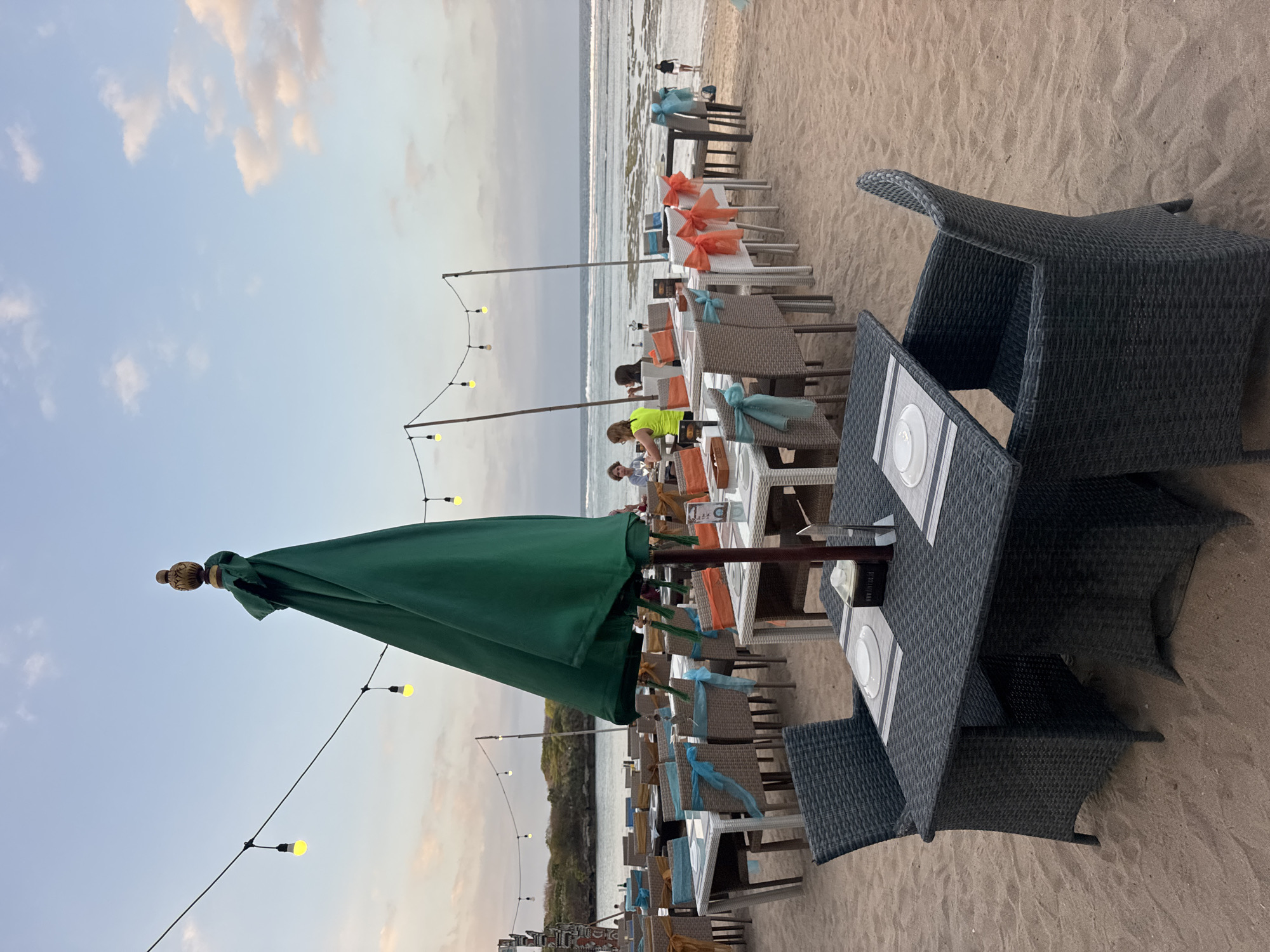 This image depicts an outdoor dining setup on a sandy beach in Kuta Selatan, Indonesia. The scene is set at dusk, with the sky displaying soft pastel colors and scattered clouds, illuminated by a string of yellow lights hung above the tables. Several tables are arranged on the sand, each set with white plates and placemats, and some chairs are adorned with decorative blue and orange ribbons. A large green umbrella is partially closed and stands prominently in the foreground. In the background, the ocean stretches towards the horizon, with a gentle shoreline and some low-lying vegetation visible on the distant landmass. A few individuals are seated at tables, engaged in dining, while others are seen walking along the beach or wading in the shallow water. The overall atmosphere suggests a tranquil and inviting dining experience by the sea.