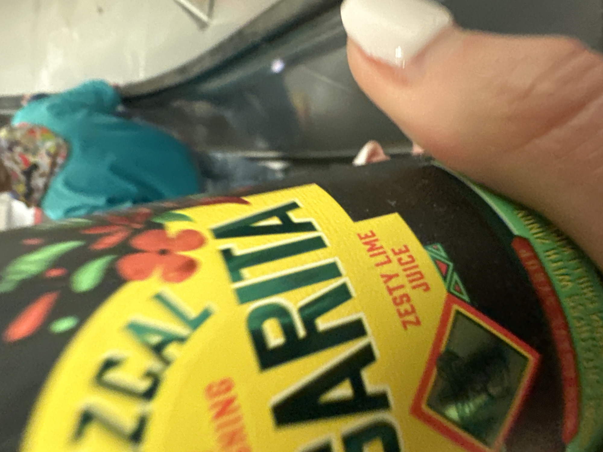 A close-up, angled photograph captures a light-skinned hand with a glossy white artificial thumb nail holding a dark-colored beverage can with a prominent yellow label. The label displays partial text including "ZCAL" and "GARITA" in green, along with "ZESTY LIME JUICE" in red, amidst red and green floral/leaf patterns. A red-bordered diamond graphic is also visible on the lower label. In the blurred background, an individual is observed from behind, standing on an escalator or moving walkway. This person wears a turquoise long-sleeved top and a multi-colored patterned scarf. The escalator features a dark handrail, and the floor of the indoor public transport environment appears light. The scene is situated within the city of London, United Kingdom.