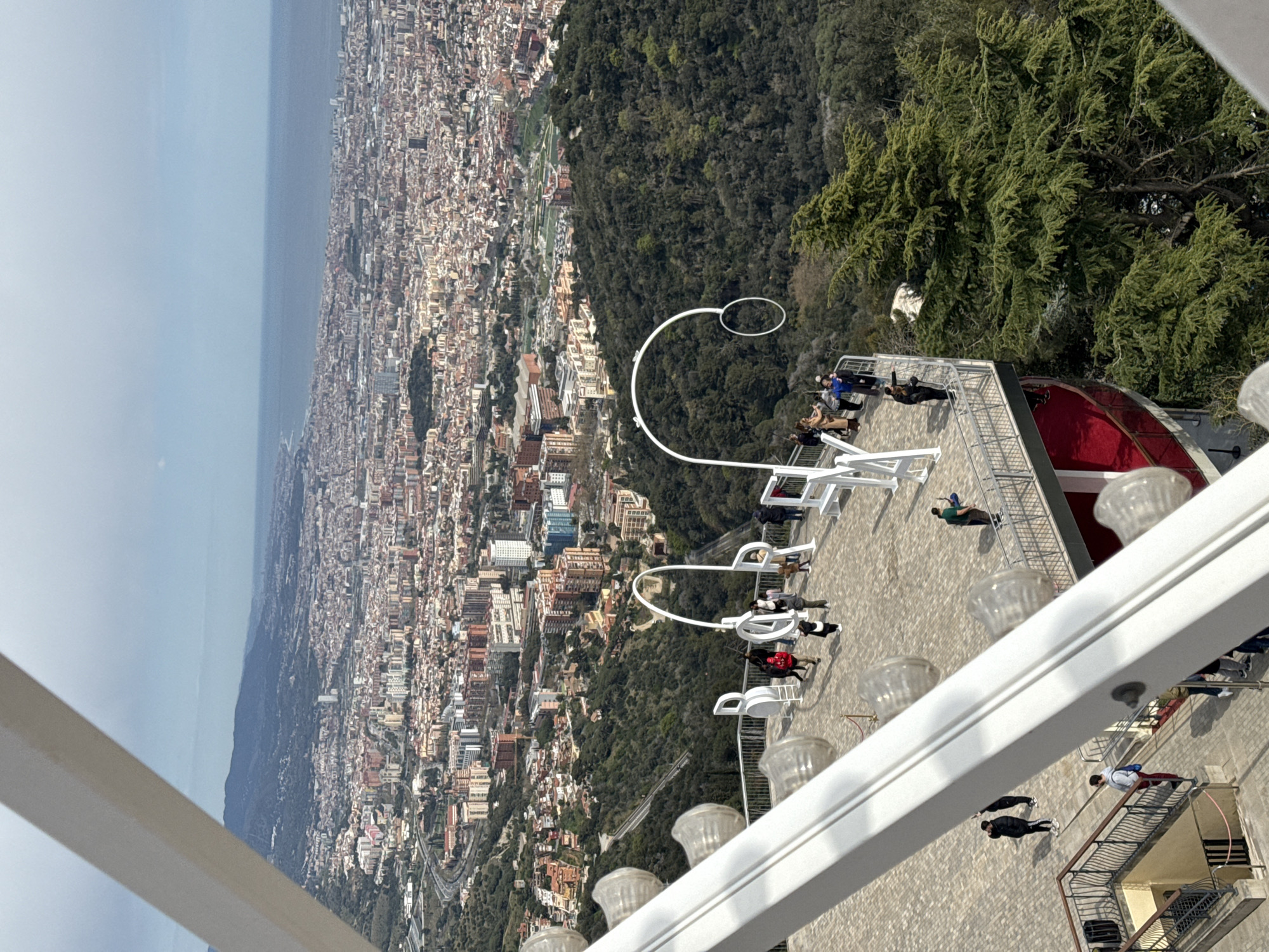 The image depicts a high-angle view of Barcelona, Spain, from what appears to be Tibidabo Mountain. The city sprawls to the left, extending towards the sea, visible in the distance. Lush green trees fill the right side of the frame, contrasting the urban landscape. On a building platform or ledge, a white artistic structure with rounded shapes is visible. Several people are standing or sitting on the platform, seemingly enjoying the view. The foreground has white architectural elements, like a frame, which obstruct parts of the view.