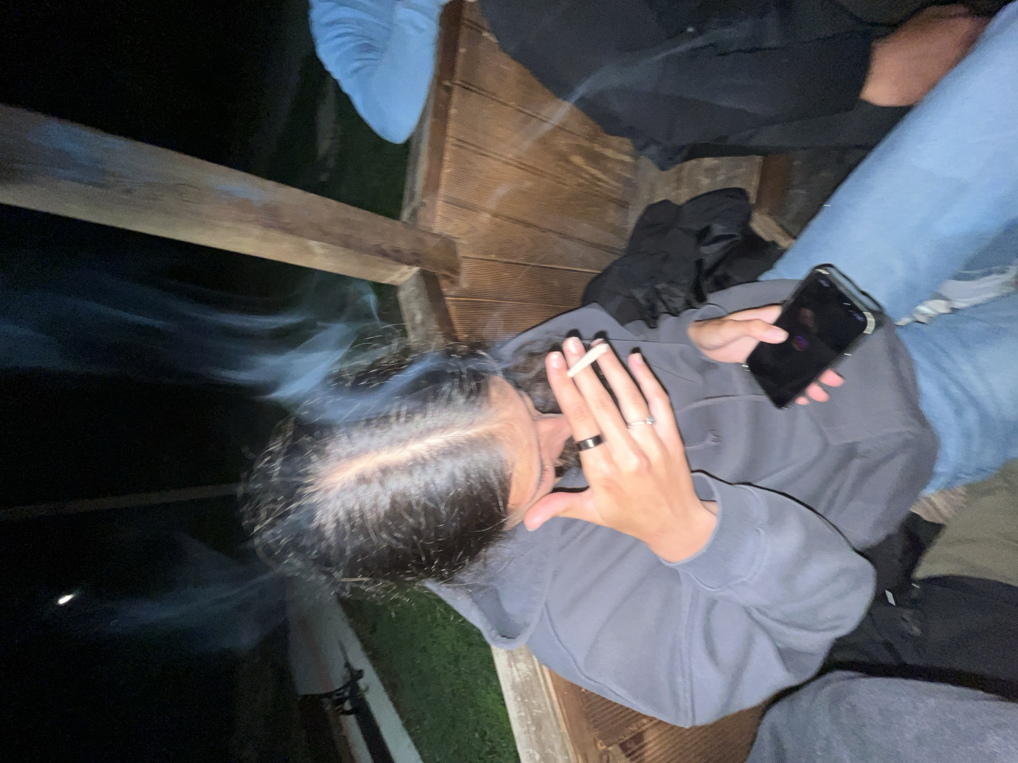 This close-up, overhead shot depicts a casual nighttime gathering on a wooden deck in Lozarevo, Bulgaria. The primary subject, wearing a grey hooded sweatshirt, is seated and engaged in two activities simultaneously: holding a lit cigarette in their right hand, with significant smoke plumes swirling around their dark hair, and holding a black smartphone in their left hand. A black ring and a silver ring are visible on their right hand.
The scene suggests a social event, as parts of at least three other individuals are visible. One person is seated to the right, wearing blue jeans, while another's blue clothing is partially seen on the upper left. Dark trousers are visible at the bottom right, indicating a fourth person. The wooden planks of the deck and a wooden railing establish the outdoor setting. The background is predominantly dark, confirming the nighttime setting. There is no visible text in the image, and emotions are not discernible from the subjects' partially obscured faces. The overall mood appears relaxed and informal.