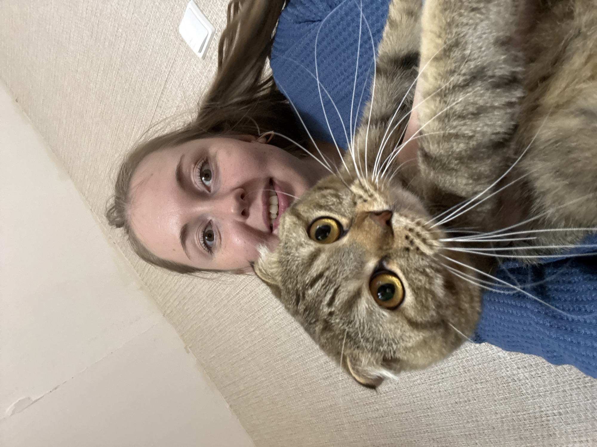 The image shows a selfie taken from a high angle, looking down at a person and a cat. The person, a young woman with long brown hair, is lying on her back on a textured, light-colored surface. She is smiling and looking at the camera. She is wearing a blue knitted garment. A cat, with brown and grey tabby markings and large yellow eyes, is positioned above the woman, also looking at the camera. The cat's face is close to the woman's, and its whiskers are prominent. The background appears to be indoors, possibly a living room or bedroom, with a plain wall visible in the upper left corner, where a white light switch or similar object is mounted. The lighting suggests it is daytime, and there are no indications of weather. The overall mood is casual and affectionate, suggesting a playful moment between the woman and her pet.