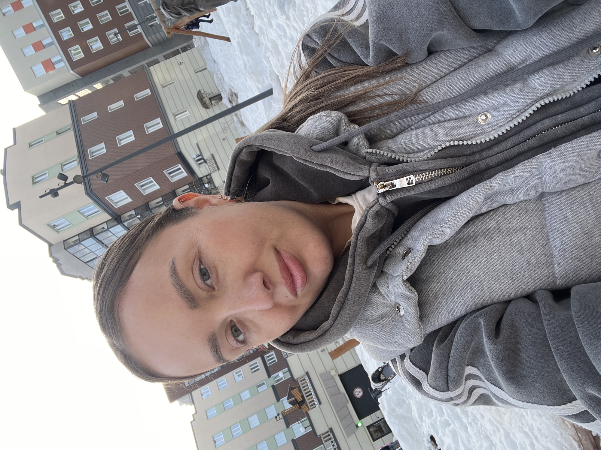 The image displays a selfie taken by a female subject outdoors in a snow-covered urban residential area within the city of Pervomaysky, Russia. The female, appearing in her 20s or 30s, is in the foreground, looking directly at the camera with a neutral facial expression. She has light brown hair pulled back and is wearing layered gray clothing: an inner light top, a dark gray hooded sweatshirt, and a lighter gray quilted vest or jacket with a visible zipper and snaps.
The background is dominated by modern multi-story residential buildings, featuring facades in beige, brown, and light green, with numerous white windows. One building has orange-colored panels near some windows. The ground is covered in undisturbed snow. A dark gray street lamp post with multiple light fixtures is visible on the left. To the right, a wooden structure consistent with playground equipment is partially visible. Further left in the midground, a child's sled is partially visible in the snow, near an indistinct human figure. A garage entrance with a "2.2M" height limit sign and a green indicator light is visible behind the subject's left shoulder. The overall environment suggests a cold, clear winter day in a developed residential complex.
