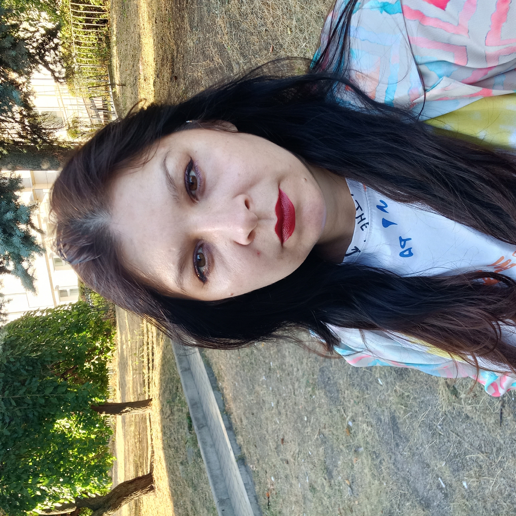 A woman with long, dark hair and a patterned jacket is taking a selfie in an outdoor urban park. The woman is looking directly at the camera with red lipstick on her lips. In the background, there are trees, dry grass, a paved walkway, and a building. The lighting suggests it is daytime, possibly late afternoon given the shadows. The location is identified as Kropyvnytskyi, Ukraine.