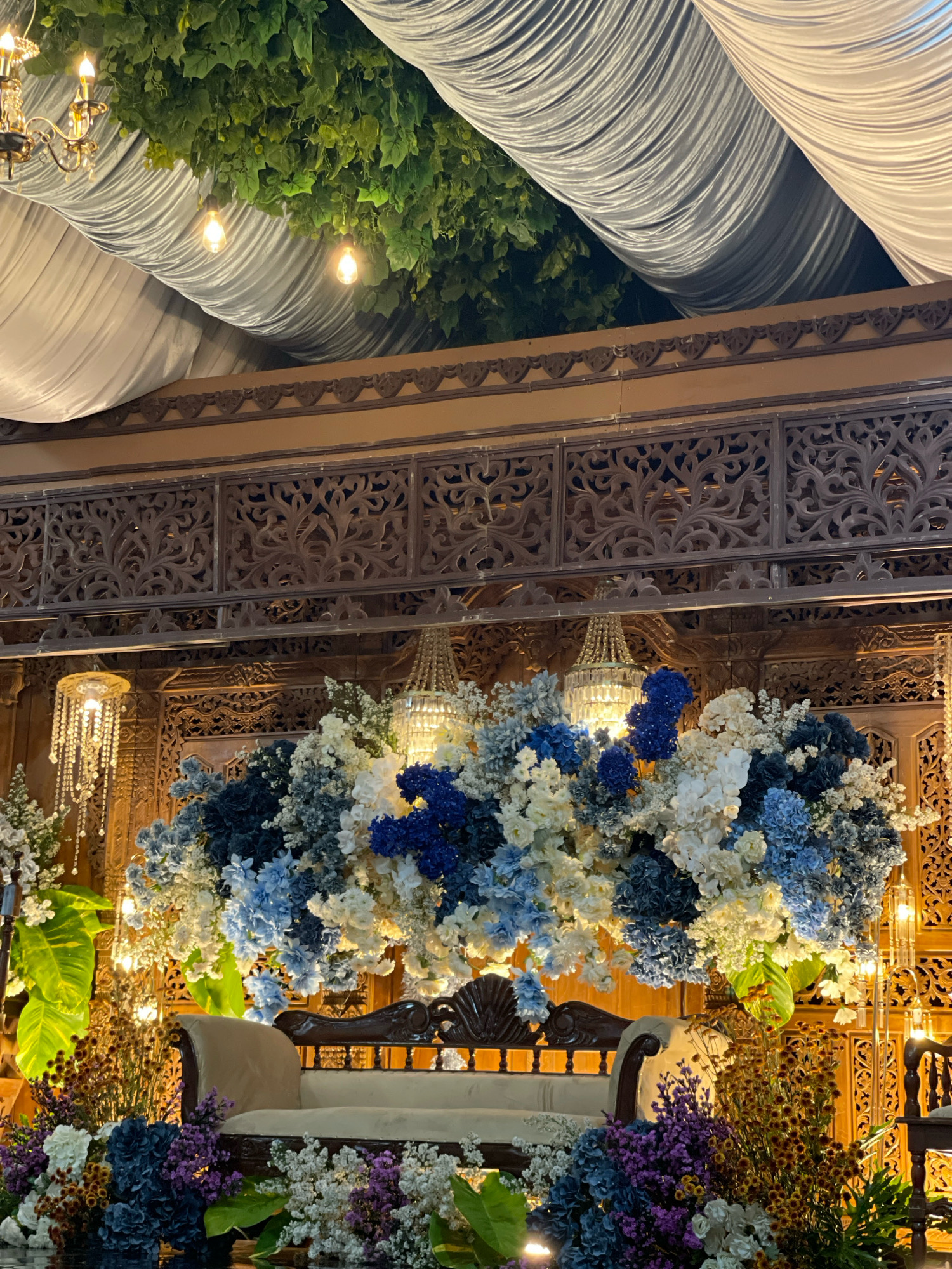 An indoor wedding stage is set up in Bagusan, Indonesia. The stage is adorned with intricately carved wooden panels and lush green foliage draped from the ceiling, interspersed with white fabric drapes. A vintage-style cream sofa sits centrally on the stage, surrounded by a voluminous floral arrangement of blue, white, and cream flowers. Crystal chandeliers hang above, casting a warm glow. The overall ambiance suggests a formal and elegant wedding ceremony.