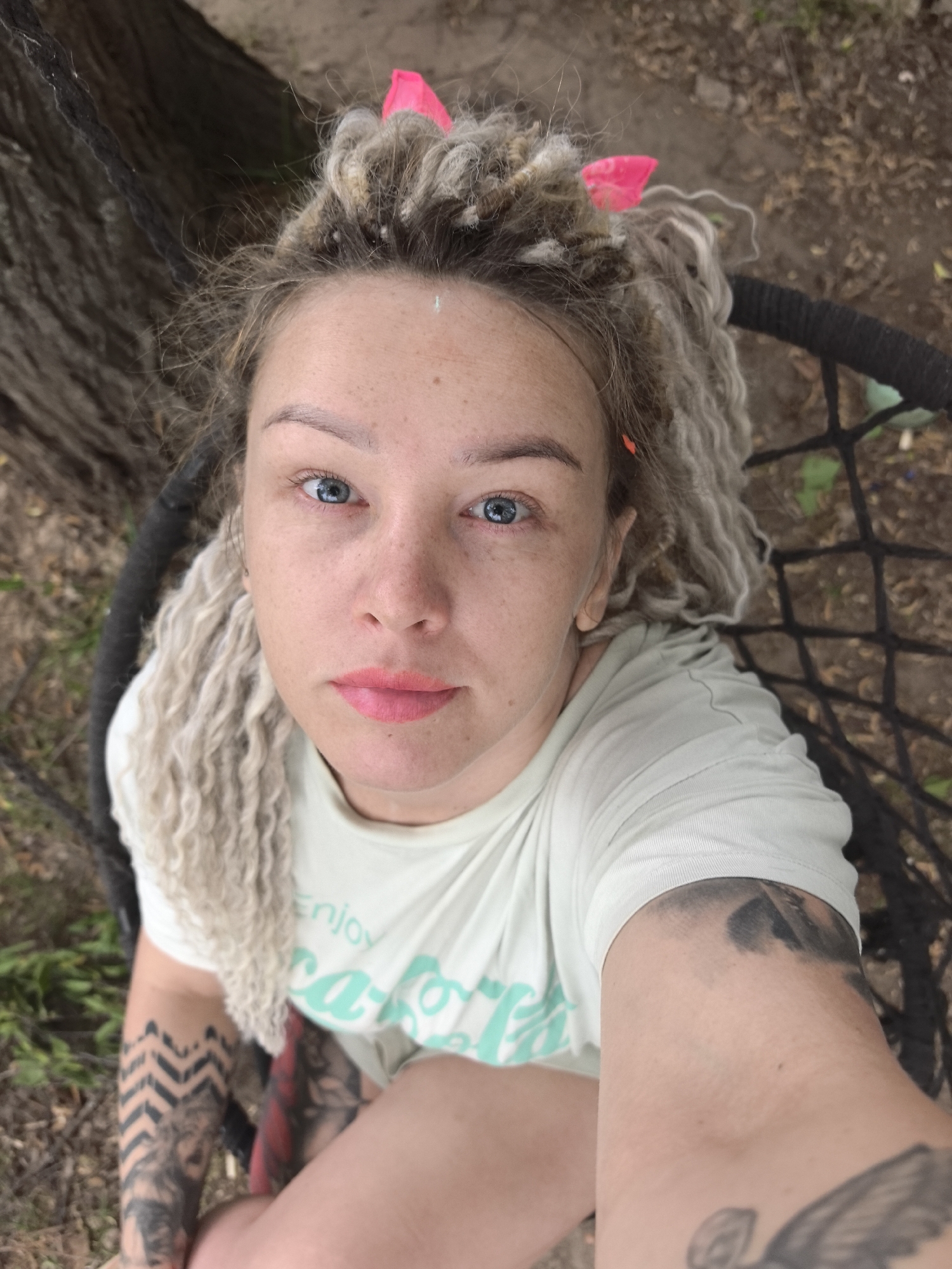 A medium eye-level selfie shows a woman in a hanging chair. Her head is tilted slightly backward to face the camera. The woman has light blonde dreadlocks with pink cat-ear hair clips on top of her head. She has fair skin and blue eyes with freckles across her nose. She is wearing light pink lipstick. She has tattoos on both arms, with a geometric design visible on her lower left arm and a bird or angel-like figure visible on her upper right arm. She is wearing a white t-shirt with a green text design. The hanging chair is a black woven type. In the background, there is a tree trunk and some grass or dirt. The setting looks like a park or outdoor area, although there is no indication of it being Noviy, Russia.