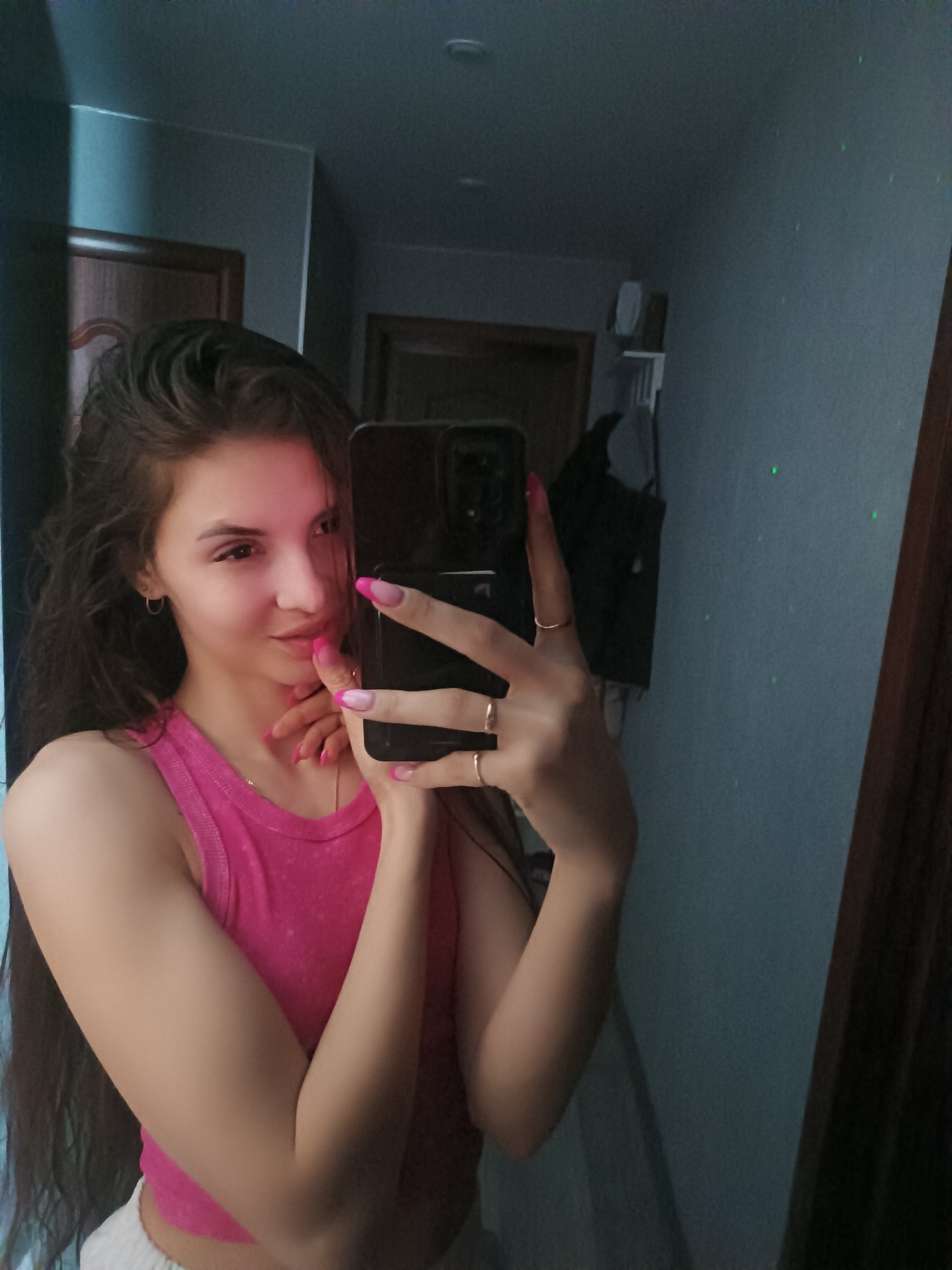 A young woman is taking a selfie in what appears to be a hallway. She is wearing a pink tank top and white shorts or pants. She has long, dark hair and is holding a black smartphone in her hand, which obscures part of her face. Her nails are painted with a pink and white design. She is wearing several rings on her fingers.
The background reveals a dimly lit hallway with doors and a wall painted in a grayish-blue color. There are scattered green laser points on the wall, and a dark garment is hanging on the wall behind her. The location of Chaikovsky, Russia is not verifiable from the image alone.