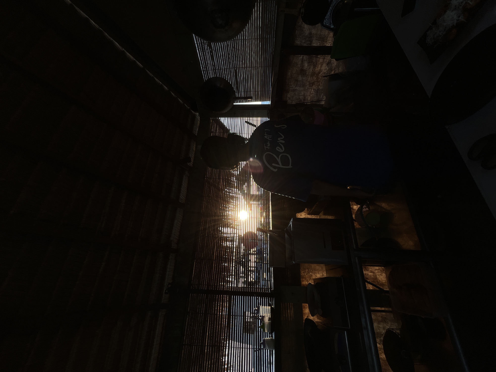 The image depicts a person standing in silhouette inside a rustic, open-air structure, likely a restaurant or food stall, in Karimunjawa, Indonesia. The setting appears to be a kitchen or preparation area. Sunlight streams through a bamboo-slatted window or door, creating lens flare and illuminating the scene with a warm glow. The person, facing away from the camera, is wearing a dark blue t-shirt with white text that reads "Fun At Ben's". Various cooking utensils and containers are visible on a counter in the foreground, cast in shadow. The overall atmosphere is serene and suggestive of an early morning or late afternoon setting.